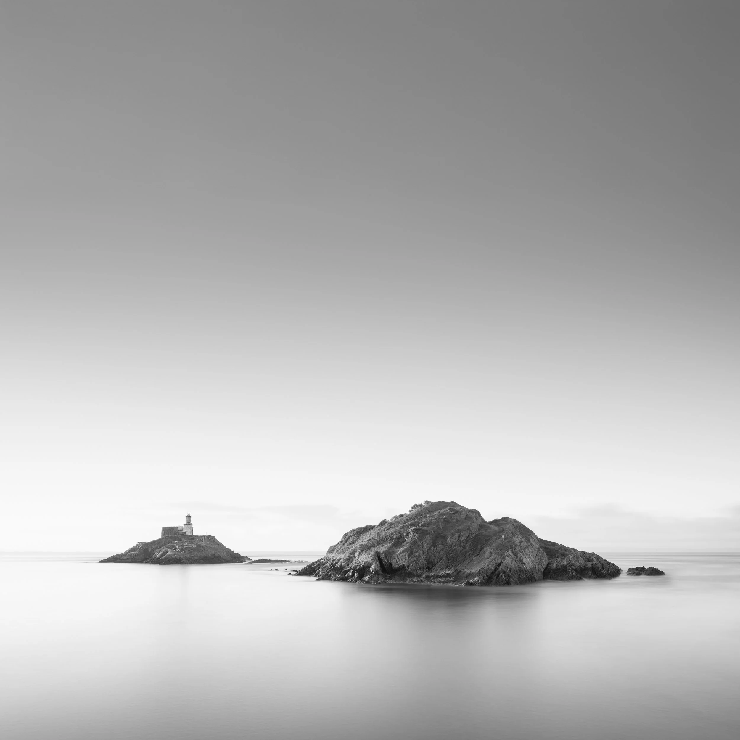 fine art Black and white photo of two rocky islands in calm water, with one island featuring mumbles lighthouse.