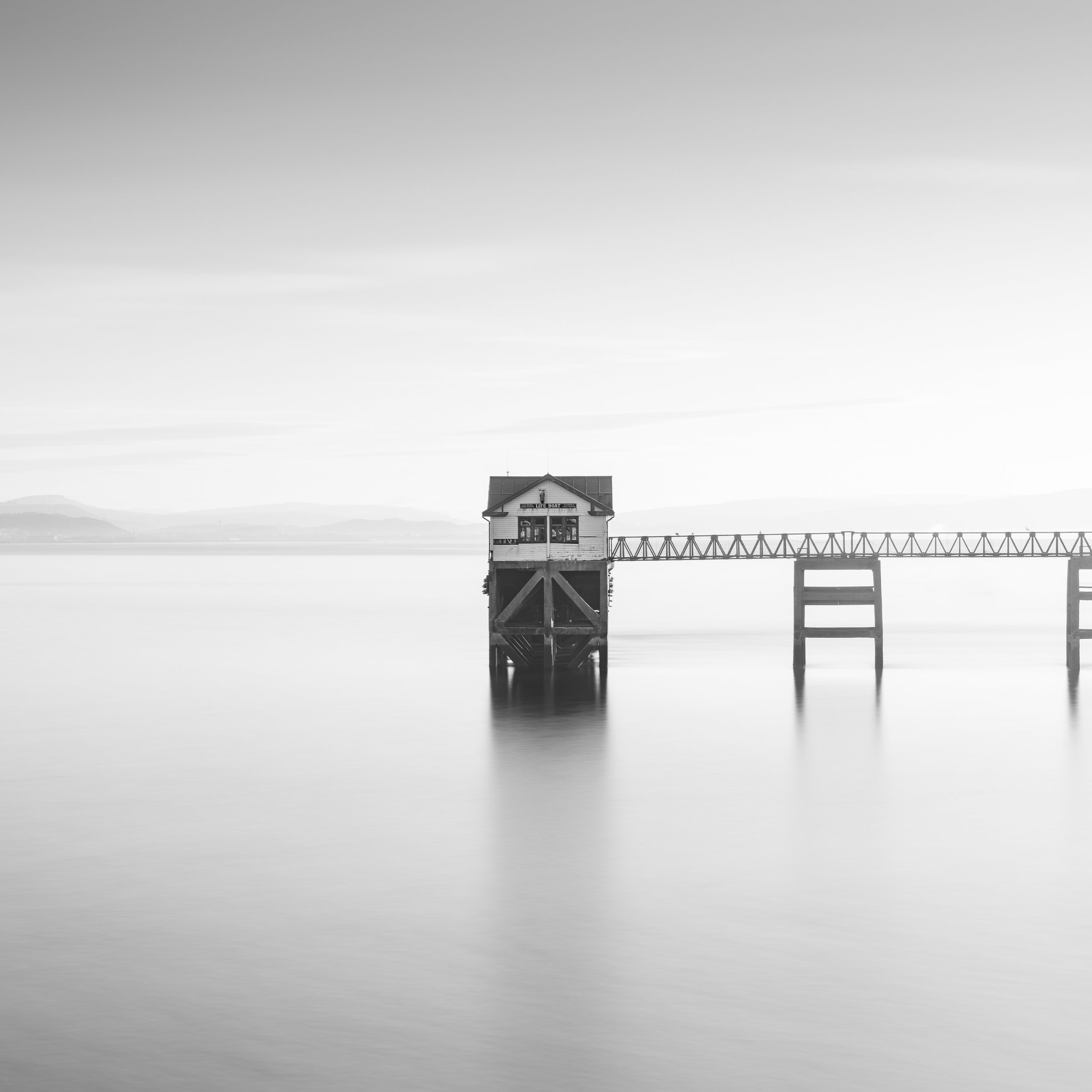 A black and white photo of mumbles lifeboat station over water connected by a pier with land visible in the background.