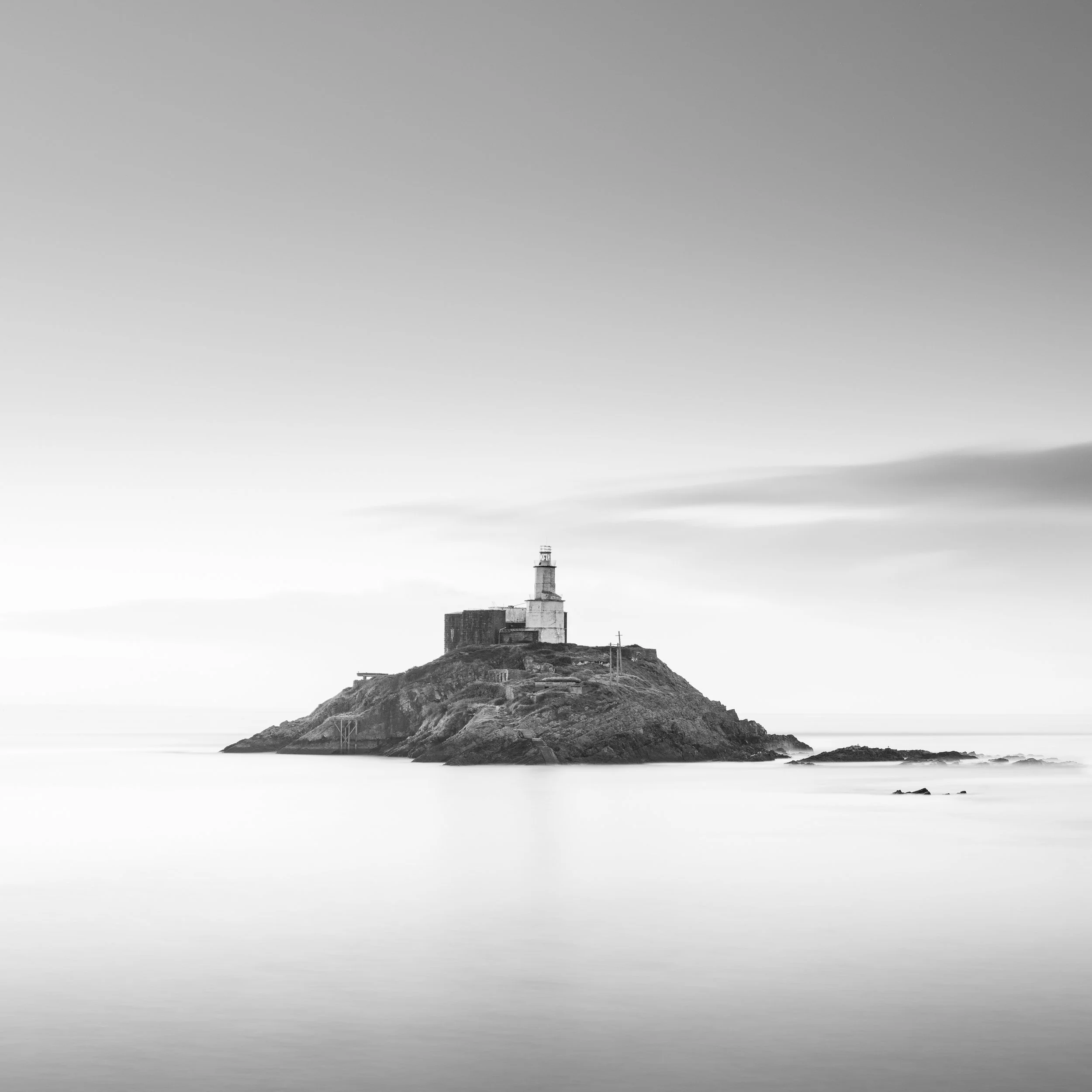 A fine art black and white photo of mumbles lighthouse on a rocky island surrounded by calm water and a cloudy sky.