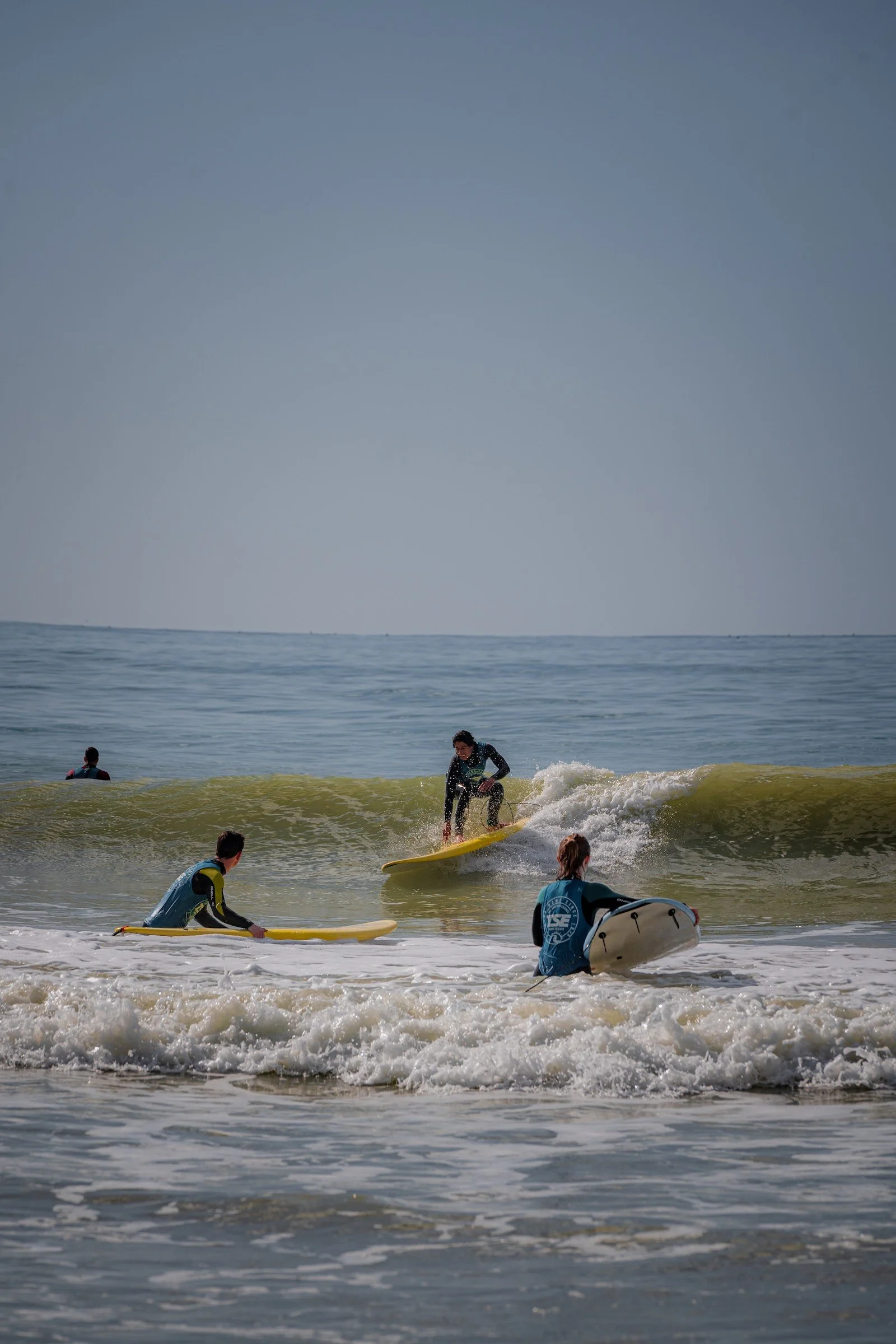 People engaging in surfing activities in the ocean, with some preparing on surfboards and others riding waves under a partly cloudy sky.
