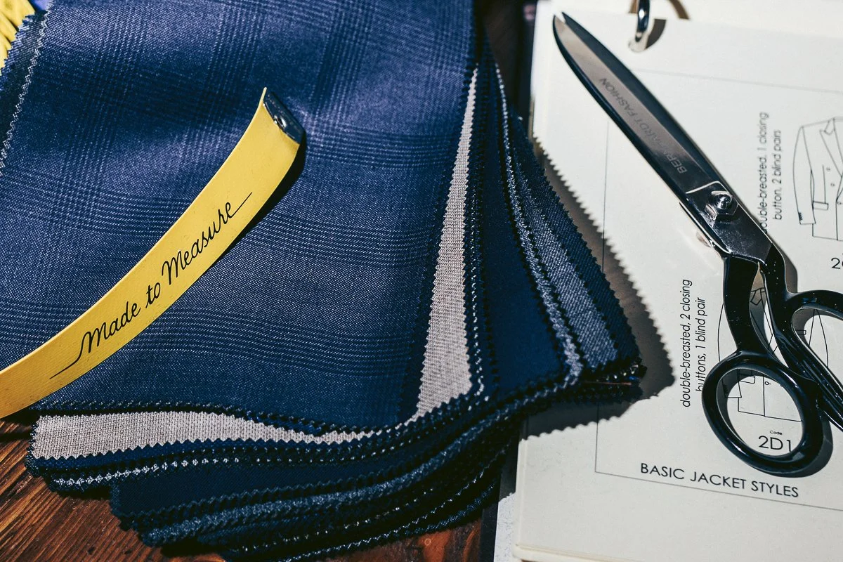 Stack of navy blue and gray fabric swatches, a yellow measuring tape reading 'Made to Measure,' a pair of black scissors, and pattern instructions for a jacket.