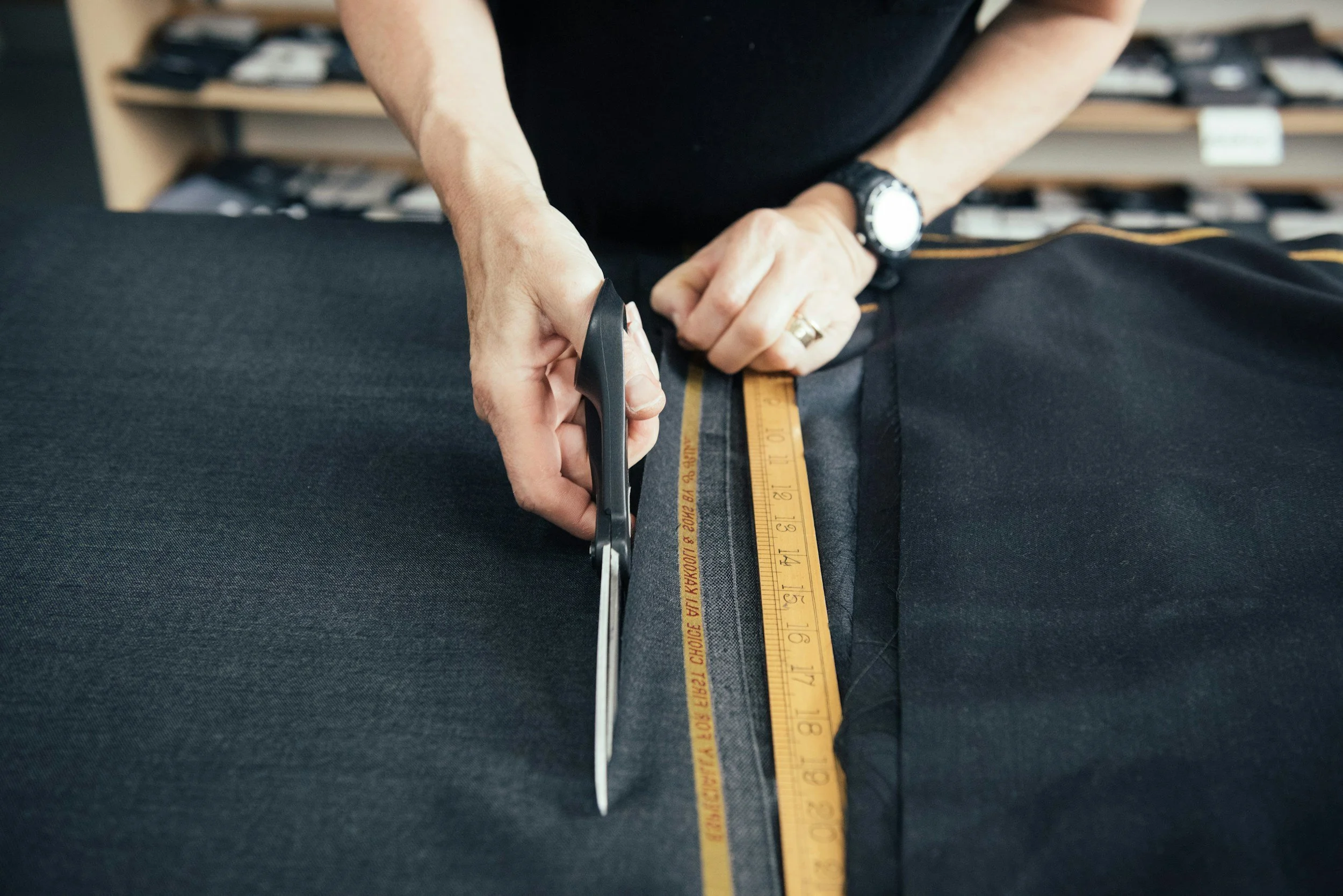 Person cutting black fabric with scissors using a yellow measuring tape to measure the fabric.