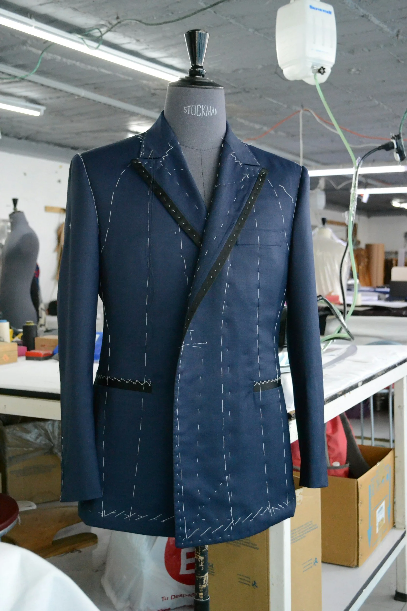 A tailored blue men's suit jacket with white chalk marks, displayed on a dressmaker's mannequin in a workshop.