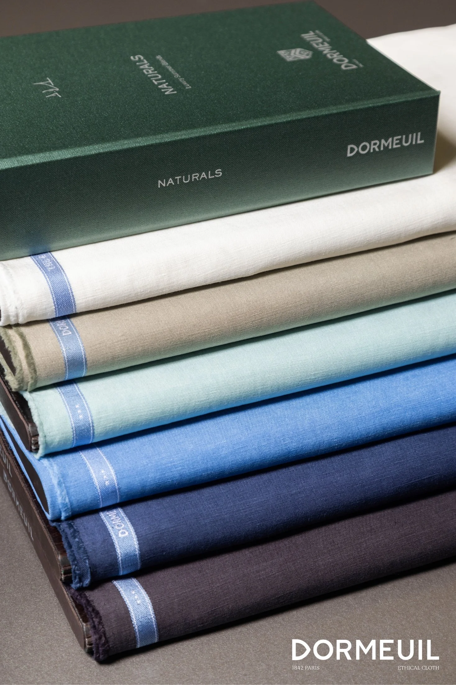 Stacked folded bed sheets in shades of beige, light green, blue, darker blue, and black with a green box labeled Dormeuil Naturals on top.