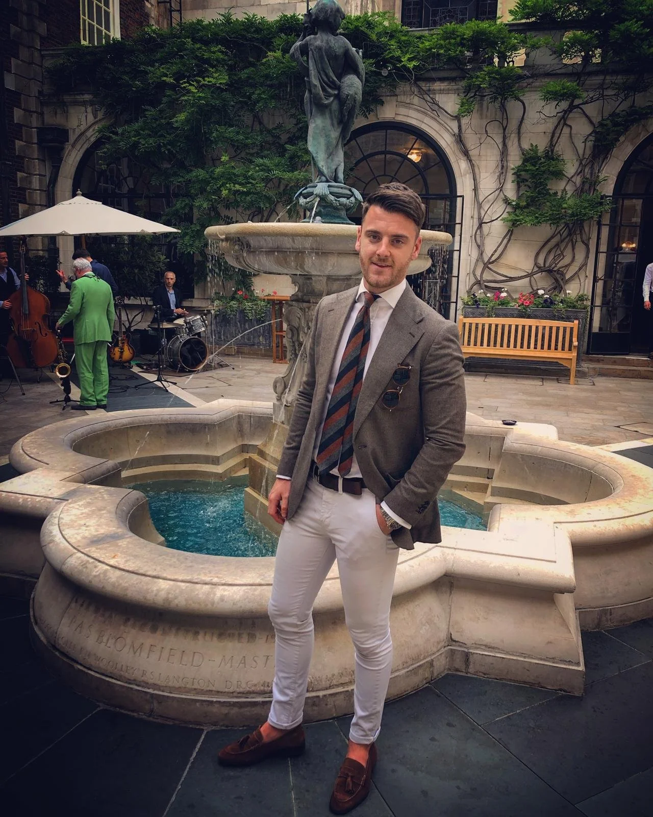 A man in a grey blazer, striped tie, white pants, and brown loafers standing in front of a fountain at an outdoor courtyard with a live jazz band playing in the background.