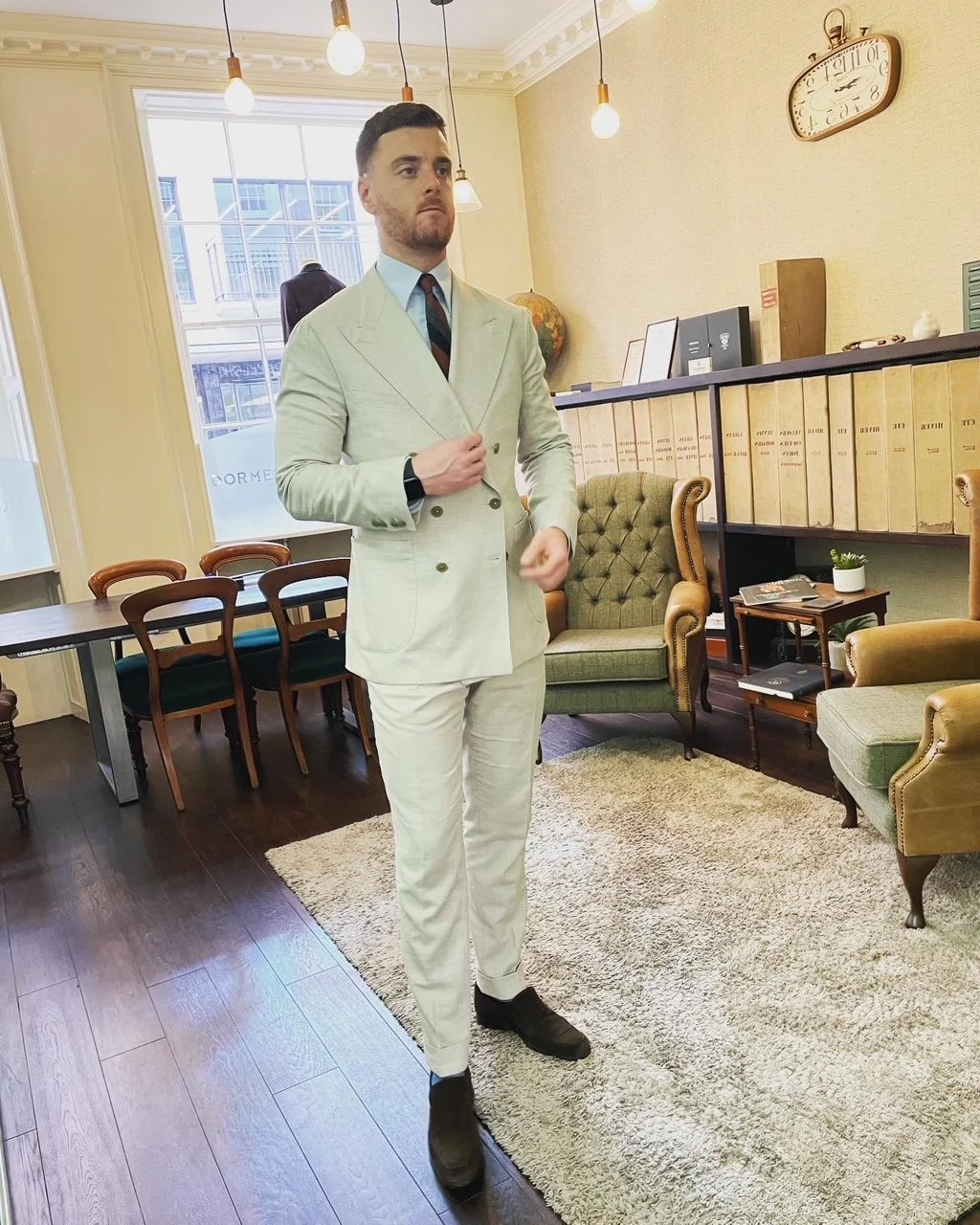 A man in a light-colored suit standing in a vintage-style office or waiting area, surrounded by chairs, bookshelves, and decorative items, with pendant lights hanging from the ceiling.