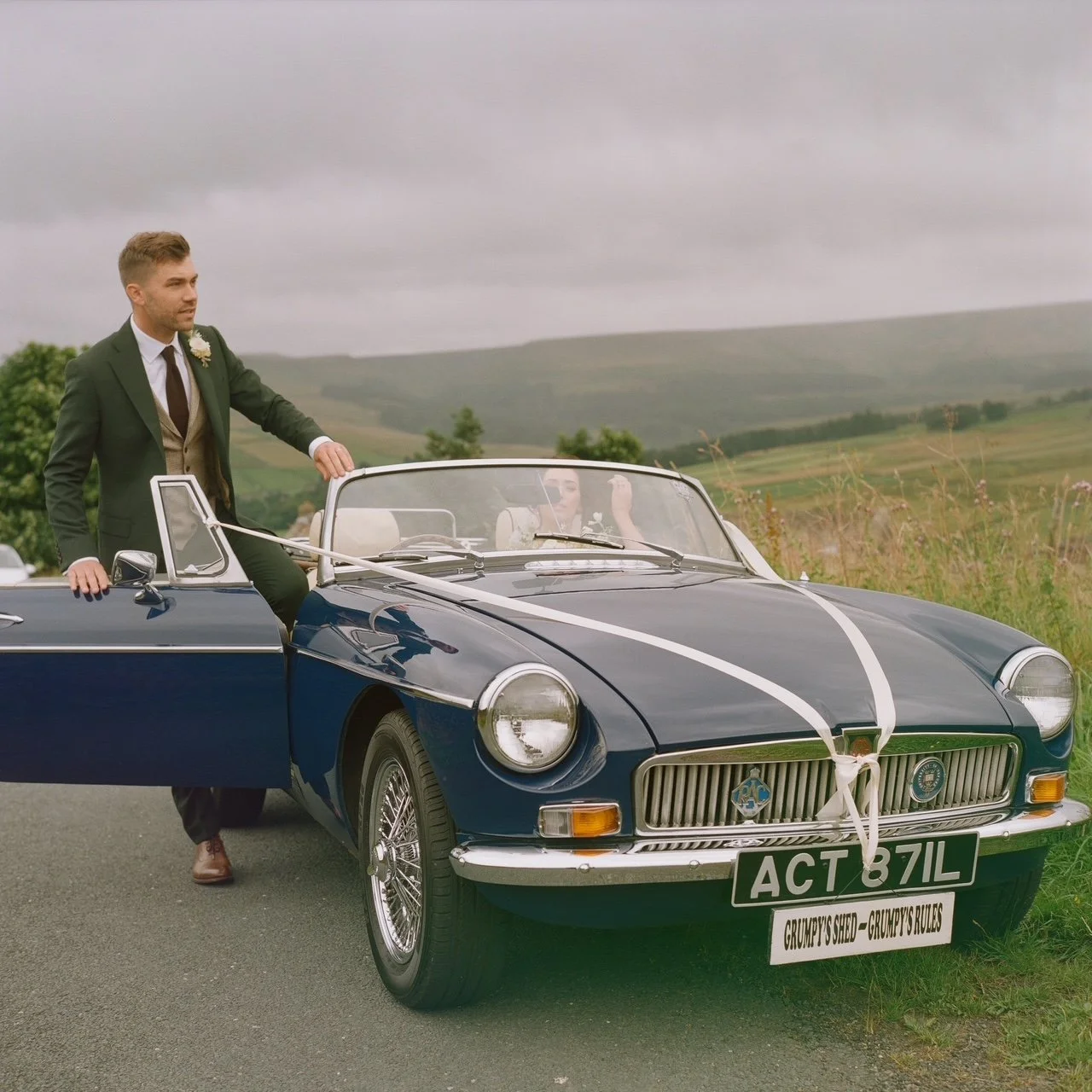 A groom standing beside a vintage blue convertible car with wedding ribbons, in a countryside setting with rolling hills and cloudy sky.