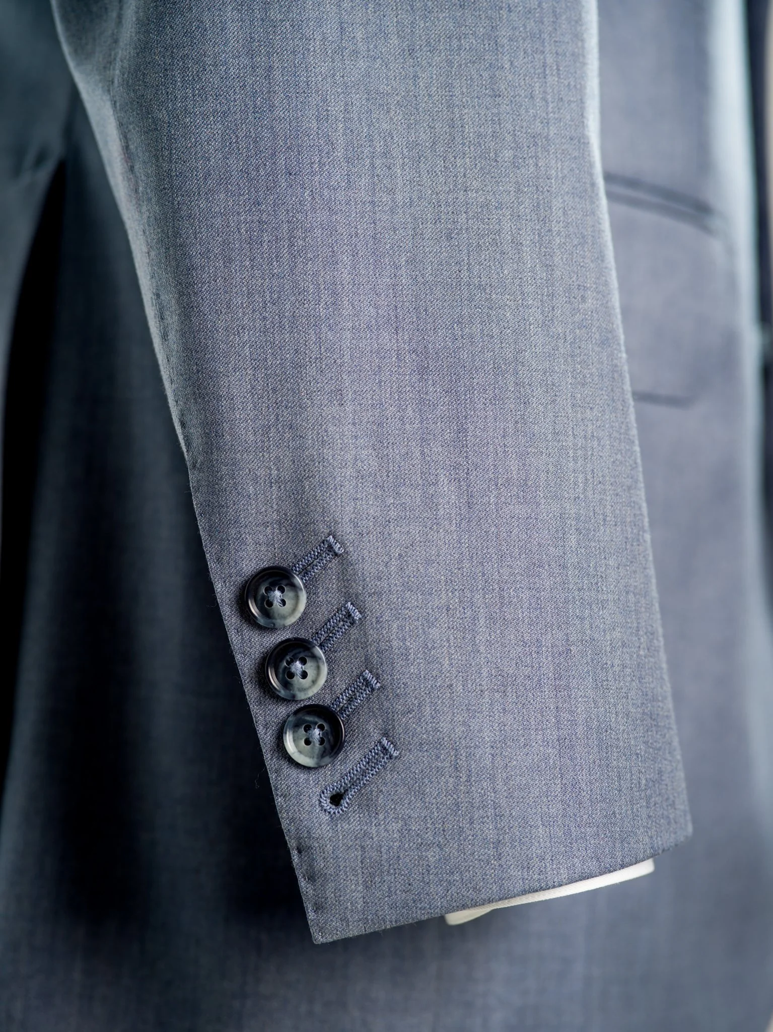 Close-up of the sleeve of a grey suit jacket showing four black buttons and buttonholes.