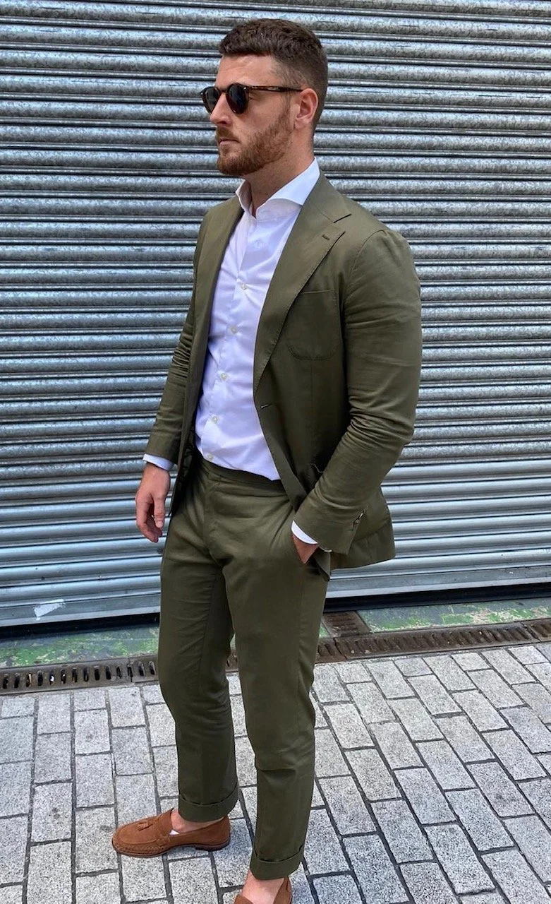 Man in olive green suit, white shirt, sunglasses, brown loafers, standing against metal garage door.