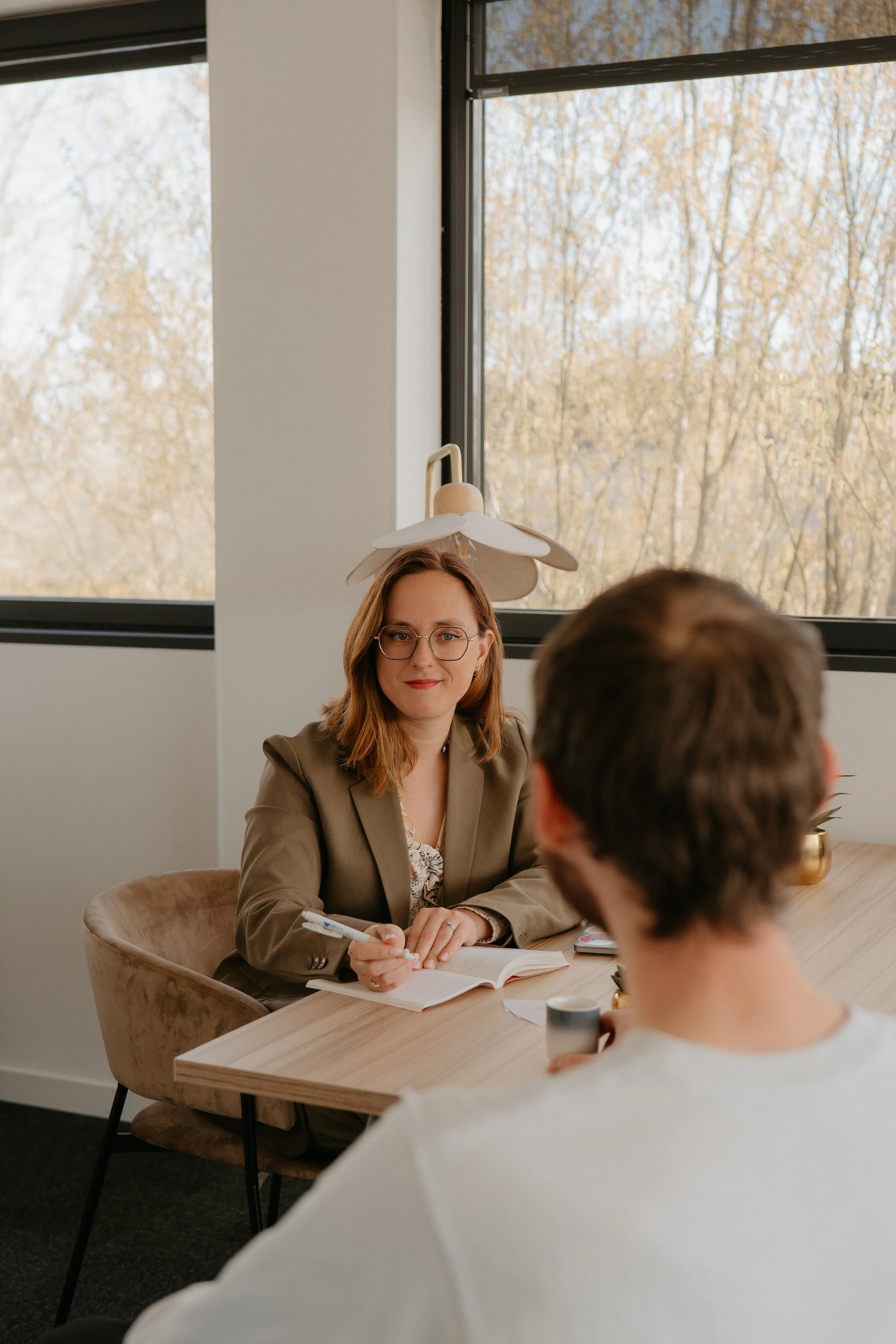 Une femme portant des lunettes et une veste kaki assise à une table, discutant avec deux autres personnes lors d'une réunion dans une salle lumineuse, avec une vue sur des arbres à l'extérieur.