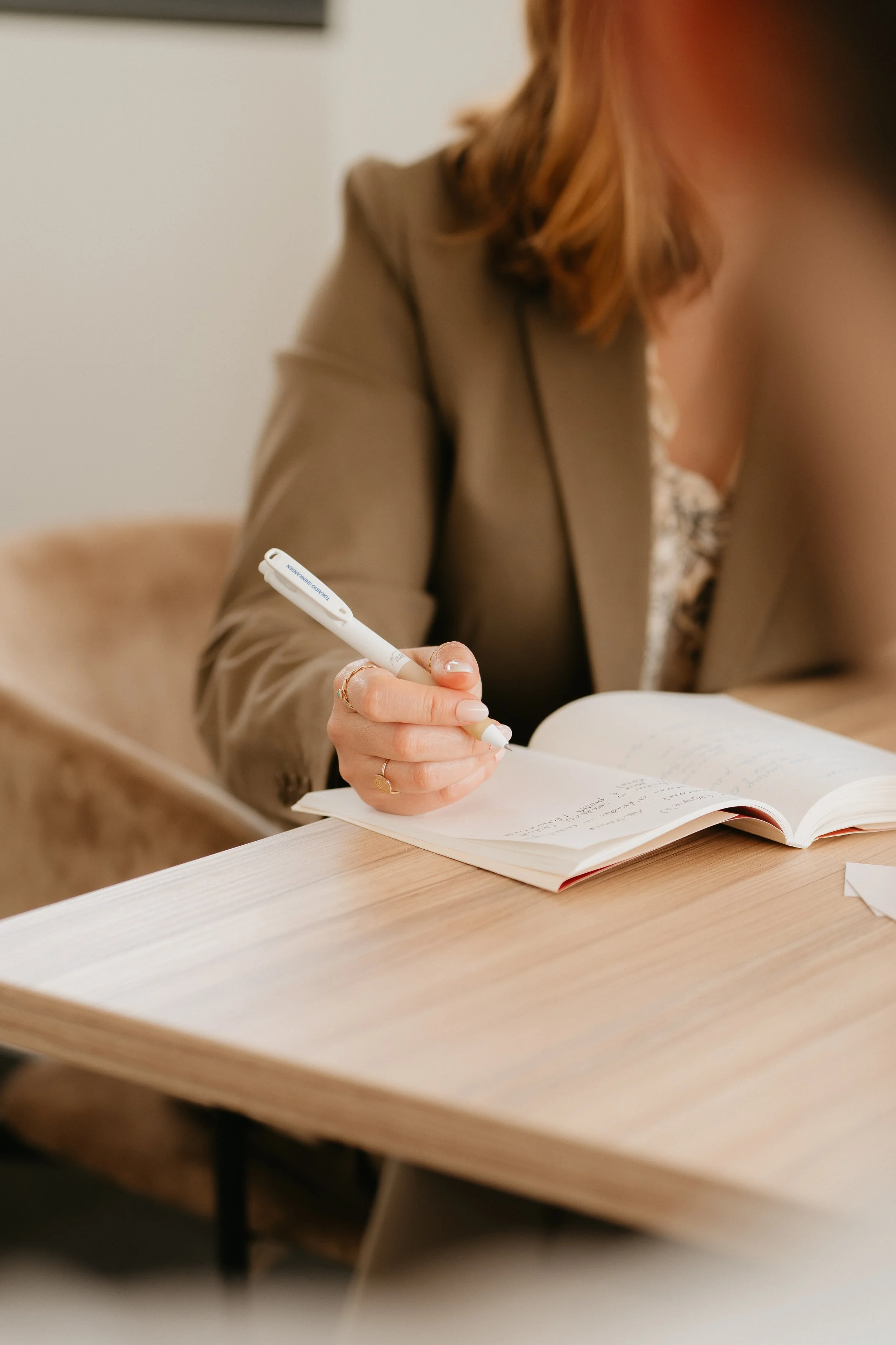 Une personne assise à une table en train d'écrire dans un carnet avec un stylo blanc. La personne porte une veste beige et a des cheveux bouclés roux. La table en bois est claire et il y a des papiers et un livre ouvert devant elle.