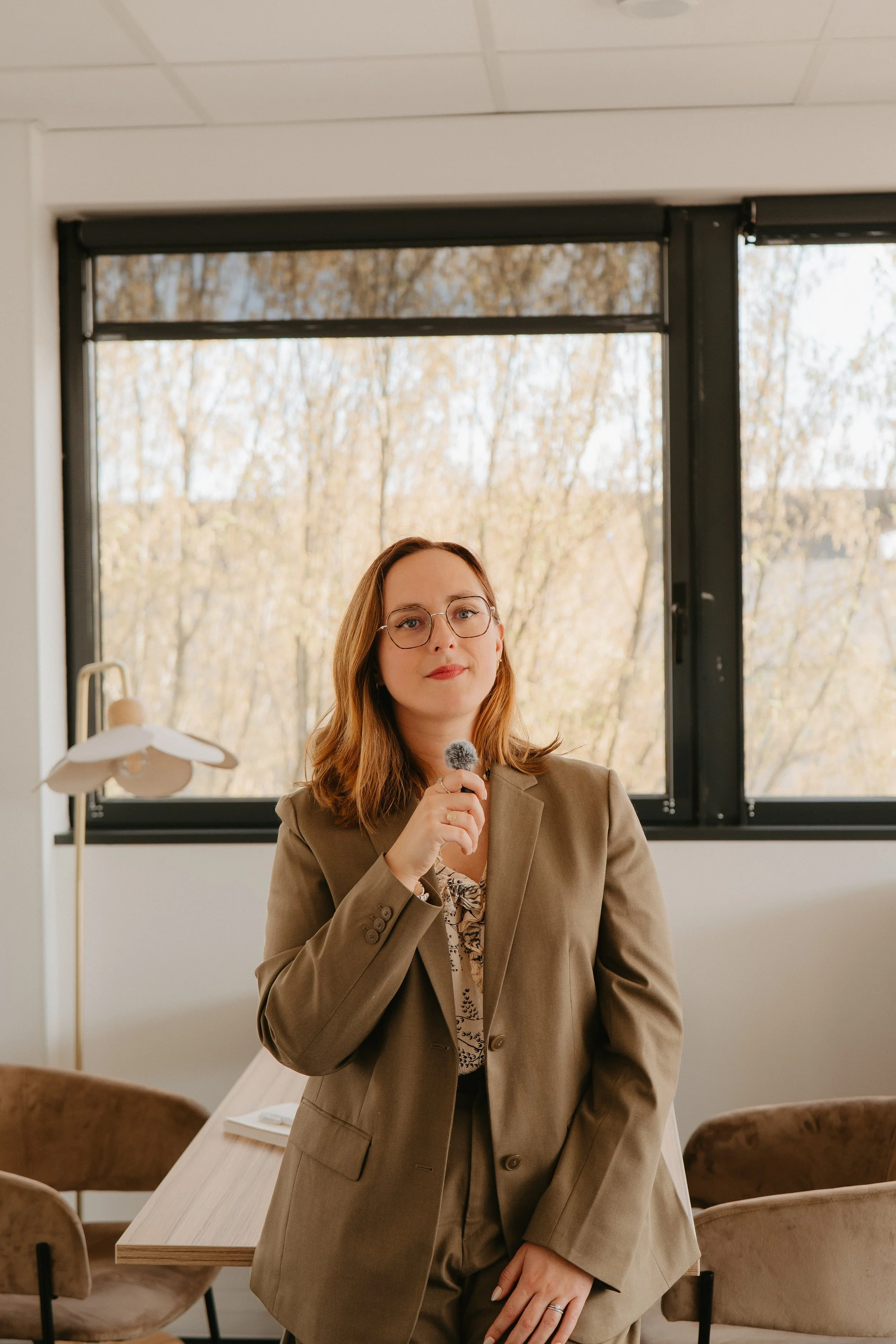 Une femme portant un tailleur beige, portant des lunettes, tenant un microphone, dans une salle avec une grande fenêtre et vue sur des arbres en automne.