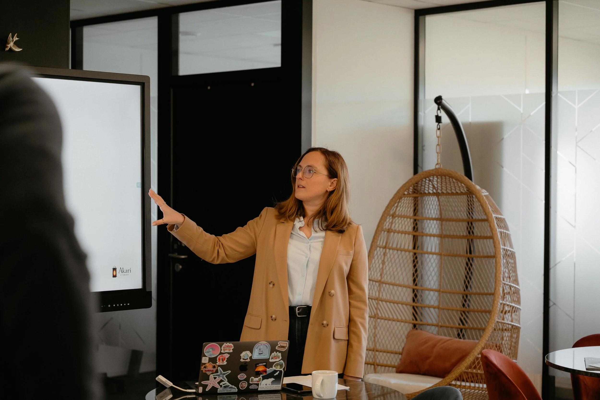Une femme avec des lunettes, portant un blazer beige, se trouve devant un écran interactif dans un bureau moderne. Il y a une chaise en rotin et une table avec plusieurs stickers et une tasse. L'ambiance est professionnelle et décontractée.