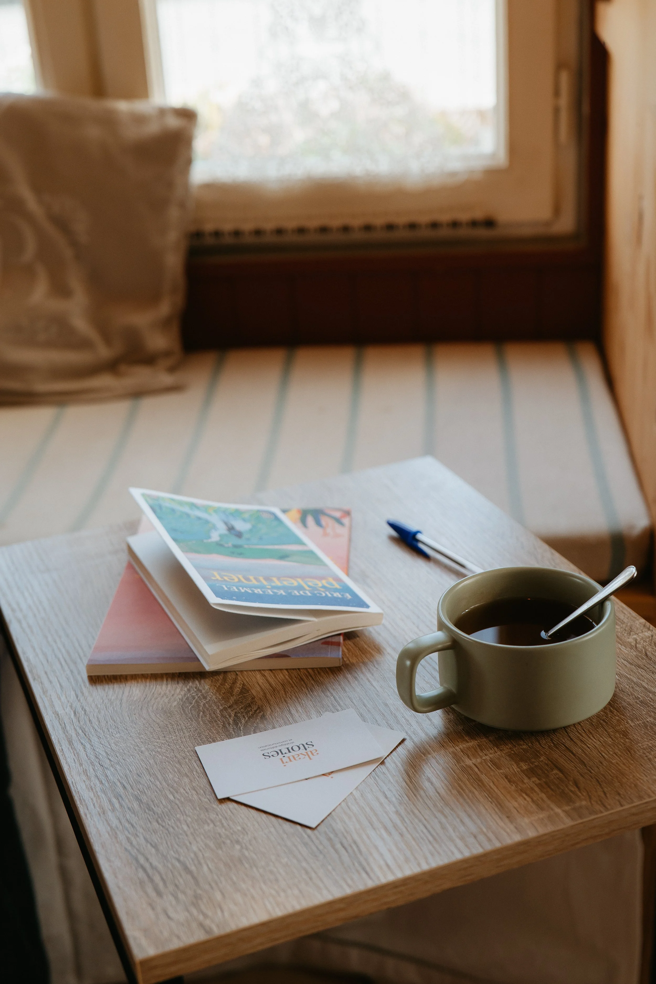 Une tasse de café noir avec une cuillère, un livre ouvert intitulé "Bild der Perfekte Design" posé sur un cahier rose, une brochure ou un guide sur la couleur, une carte de visite ou un flyer intitulé "Aktion Solutions", sur une table en bois, près d'une fenêtre qui laisse entrer de la lumière naturelle.