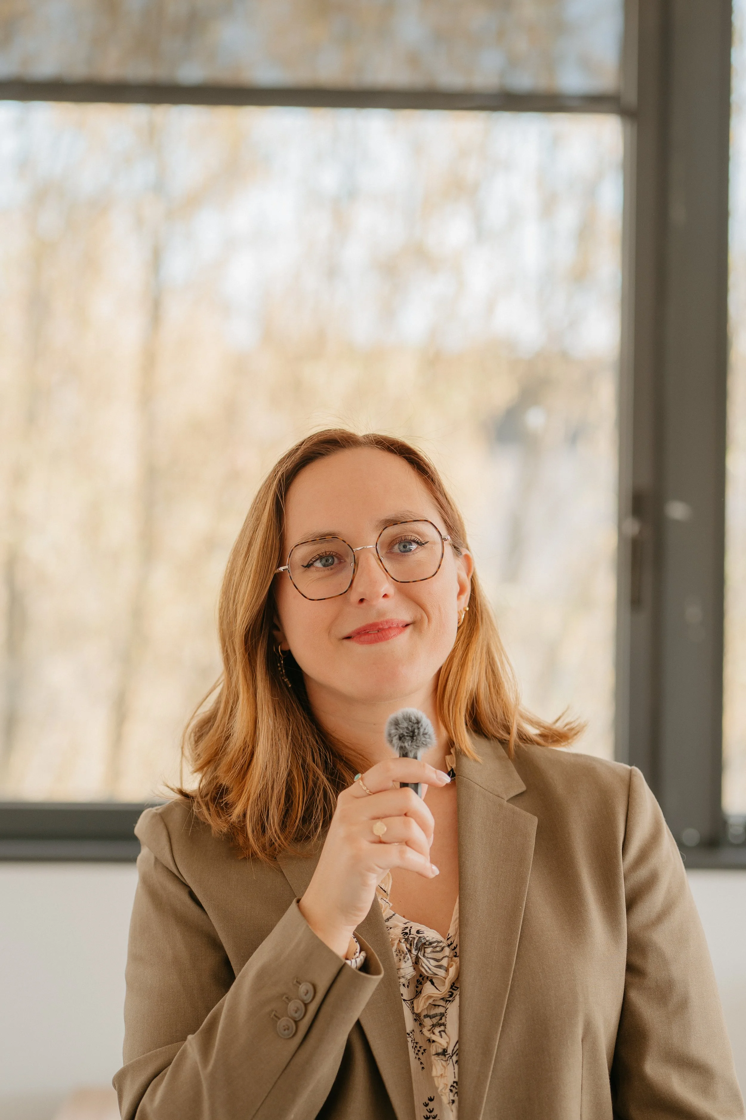 Une femme avec des cheveux roux, portant des lunettes rondes, un tailleur beige et une blouse à motifs, tenant un micro à main, devant une grande fenêtre en arrière-plan.