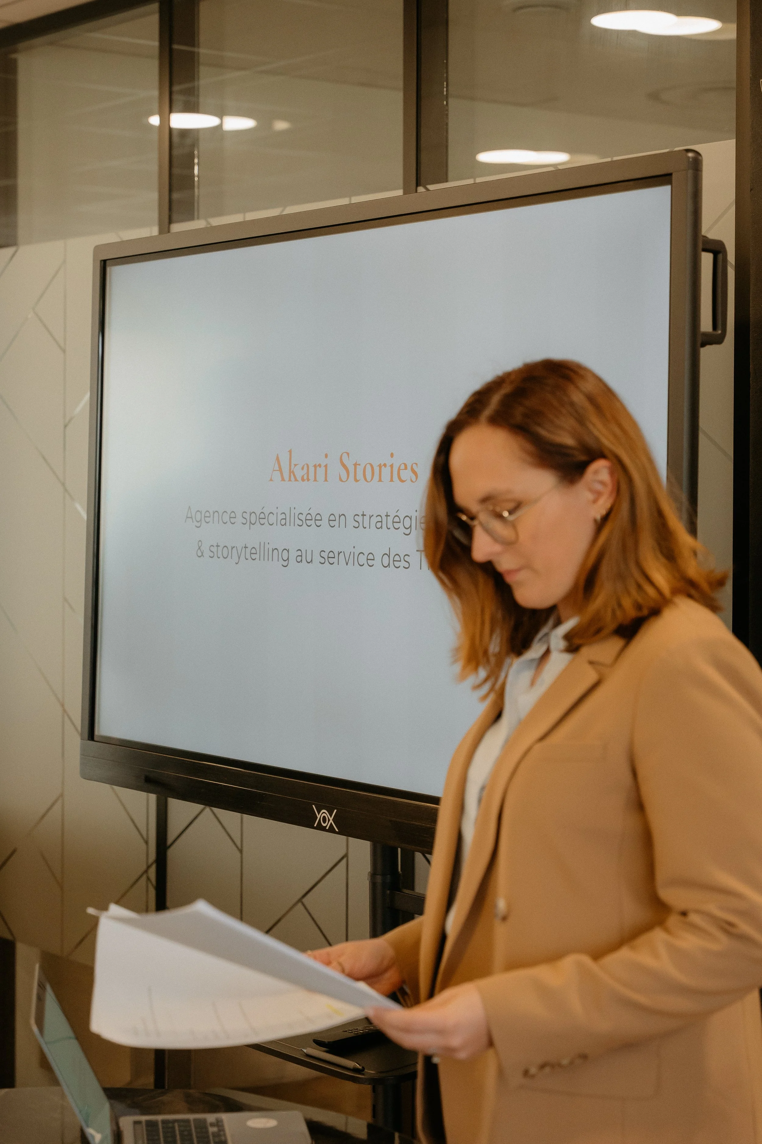 Une femme portant un blazer beige lit un document devant un écran affichant 'Akari Stories' et une description en français. Elle se trouve probablement dans un environnement professionnel ou un événement.