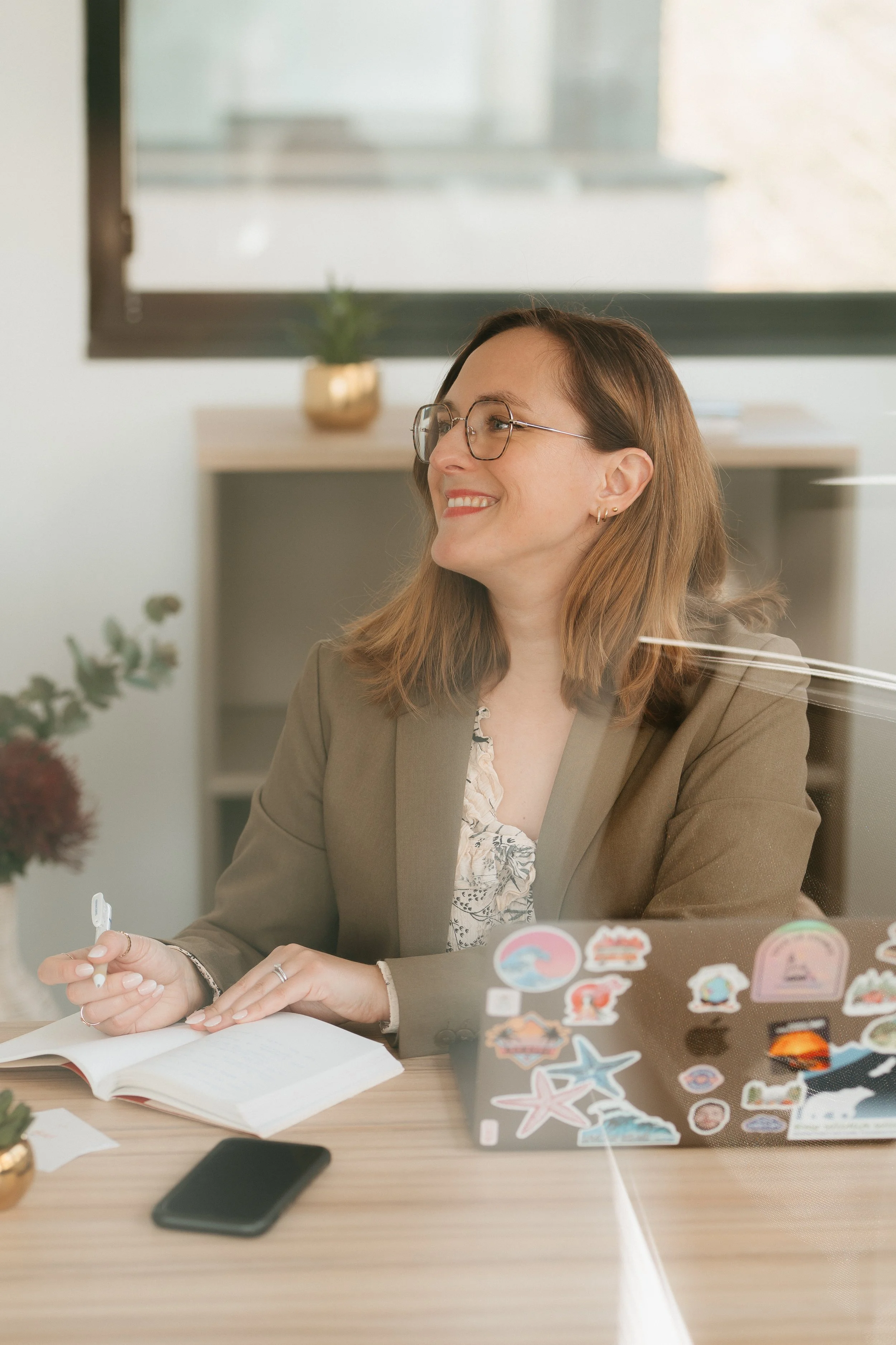 Une femme souriante avec des lunettes assise à une table dans un bureau, tenant un stylo et ayant un ordinateur portable décoré de stickers colorés.
