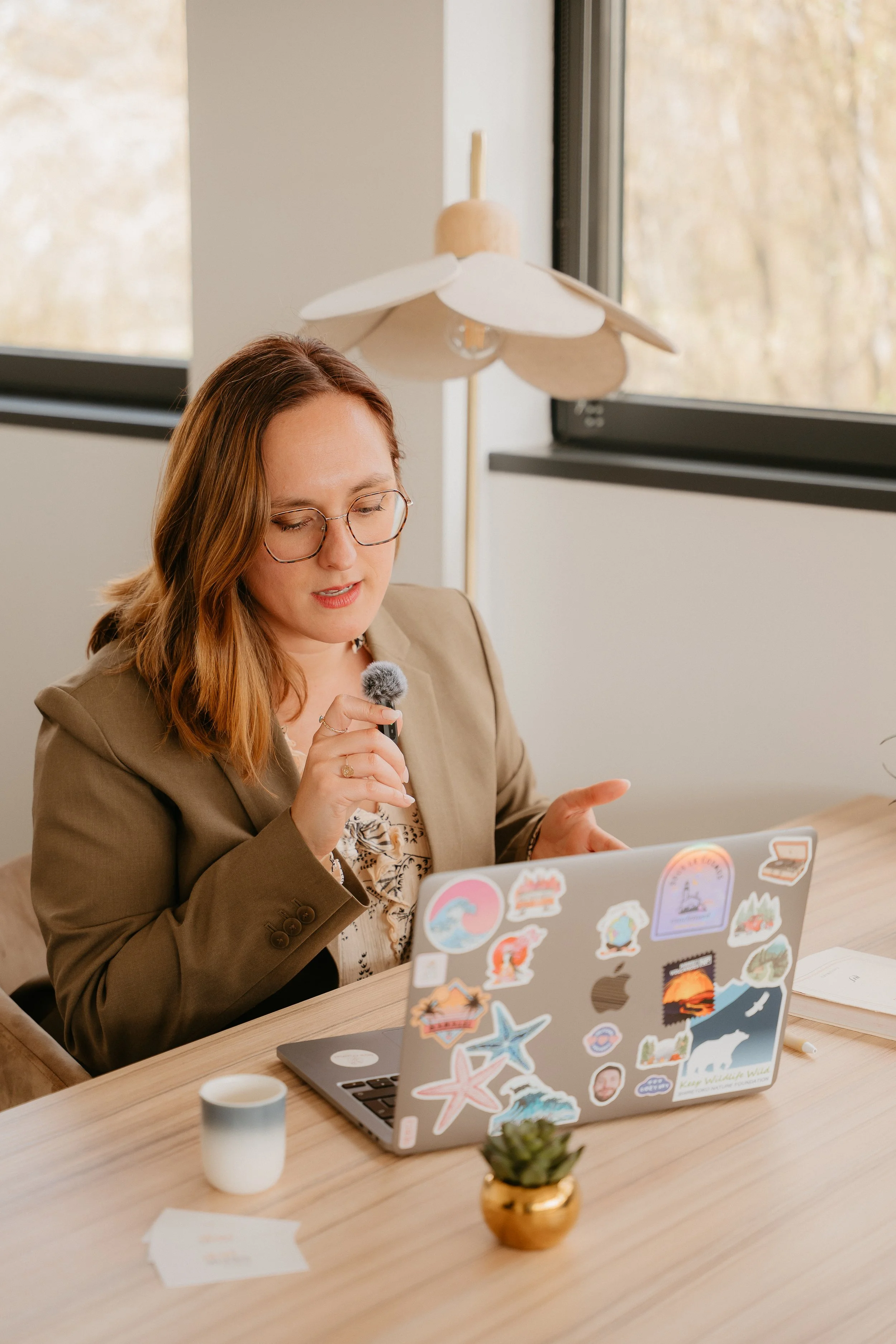 Une femme portant des lunettes, assise à une table, utilisant un ordinateur portable décoré avec des autocollants, dans un environnement lumineux avec une grande fenêtre et une lampe au plafond.