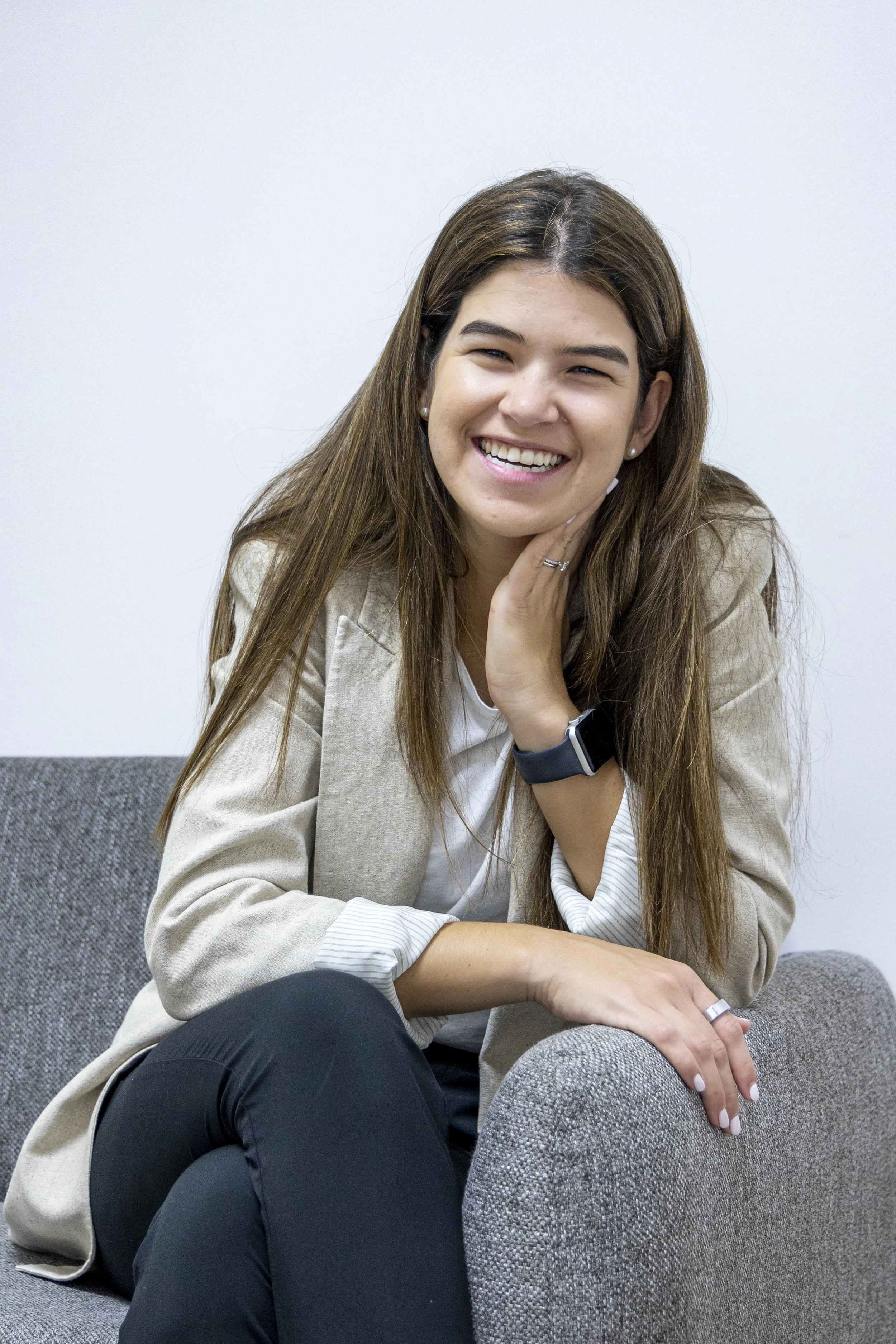 A smiling woman with long brown hair, wearing a beige blazer, white shirt, and black pants, sitting on a gray couch against a white wall.