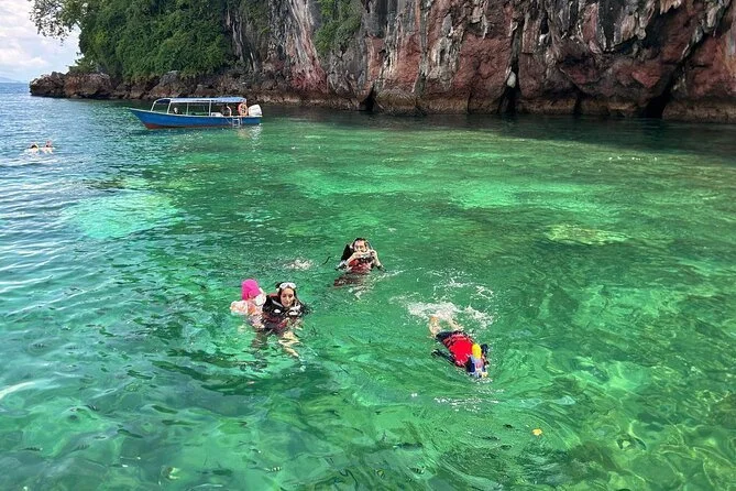 Three children swimming in clear, greenish water near rocky cliffs, with a boat anchored nearby.