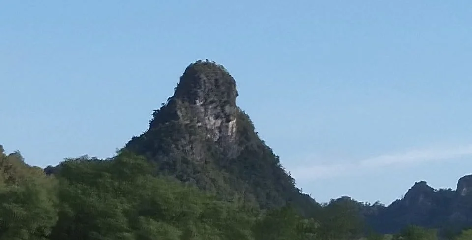 A mountain with a steep, rocky peak surrounded by green trees under a clear blue sky.