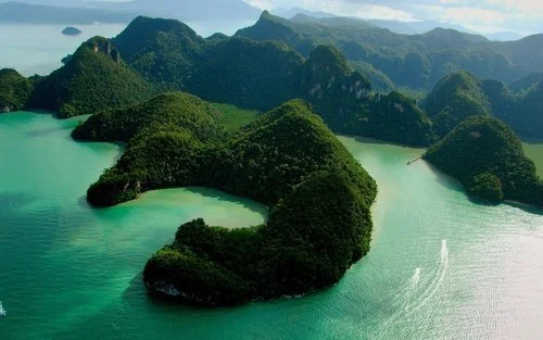 Several lush green islands with steep hills surrounded by turquoise water, with a boat creating a wake in the water.