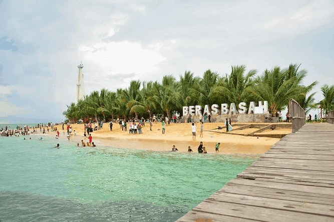 Beach with turquoise water, people swimming and walking along the shore, a group gathering near palm trees, a large white sign that reads 'BERBASAH,' and a wooden pier on the right side.