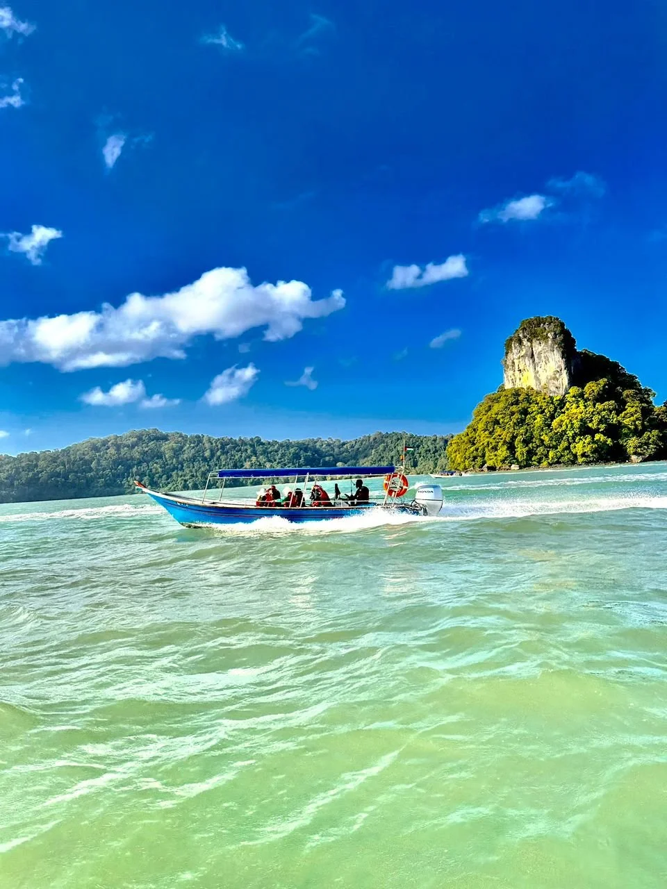 Speedboat on turquoise water near green island with rocky cliffs and a bright blue sky with scattered clouds.