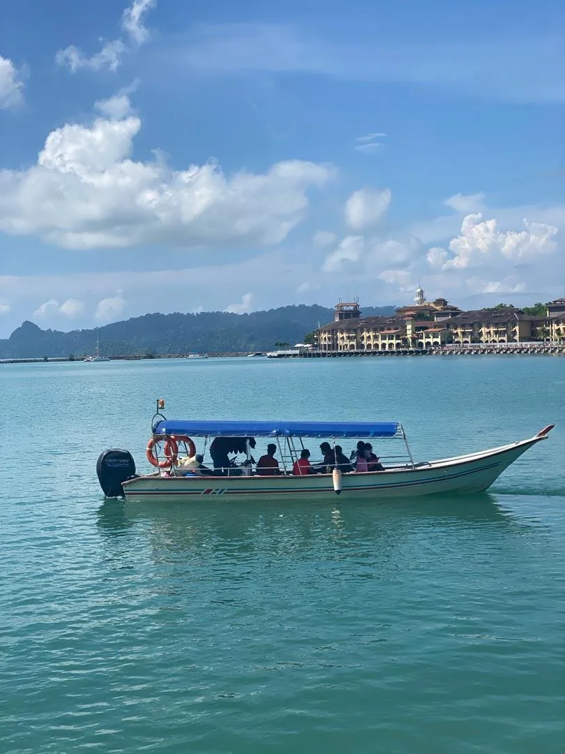 A boat with a blue canopy carrying several passengers on calm turquoise water near a coastline with buildings and mountains in the background under a partly cloudy sky.