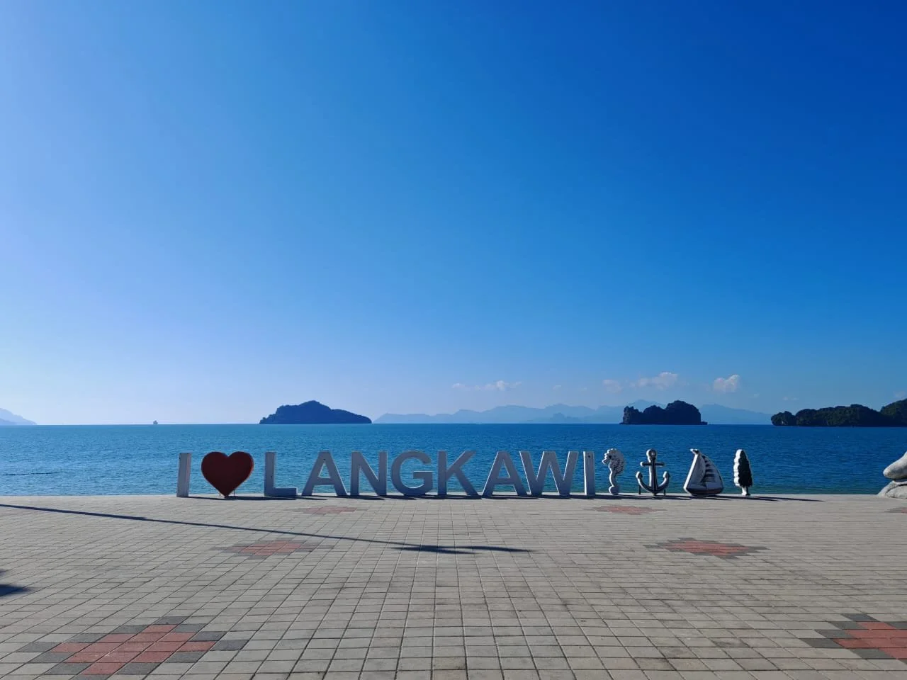 A large sign spelling 'I ❤️ LANGKAWI' with nautical and island-themed decorations, situated on a paved promenade by the ocean under a clear blue sky.