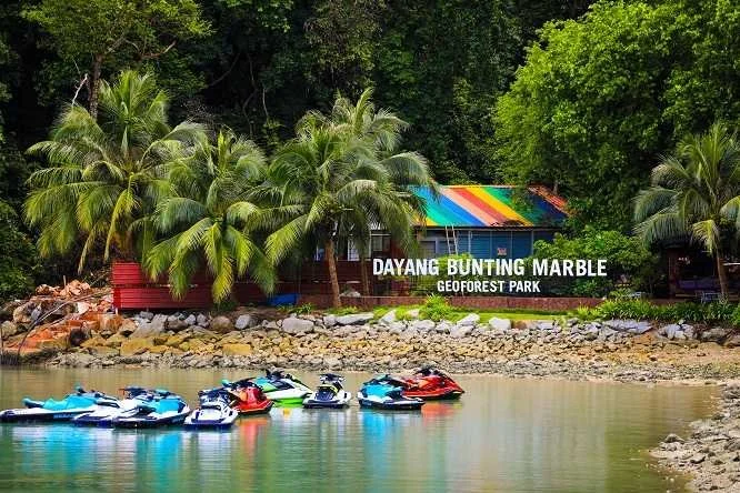 Kayaks and jet skis floating on a river near palm trees and a colorful building with a rainbow roof at Dayang Bunting Marble Geoforest Park.