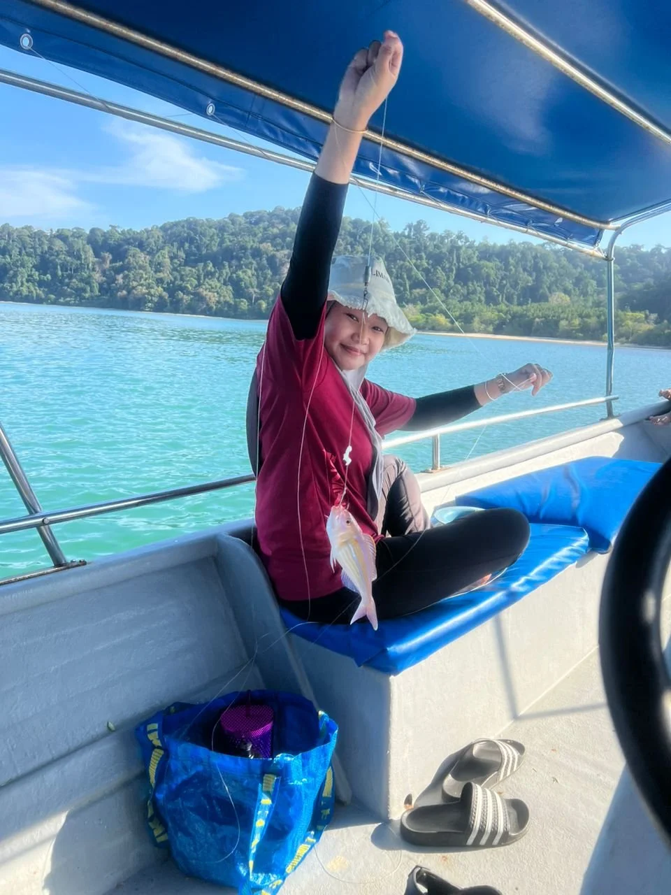 A girl sitting on a boat holding up a fishing line with a fish she caught, smiling. She is wearing a hat, a red shirt, and black leggings, with slippers on the floor. The background features a body of water and lush green hills.