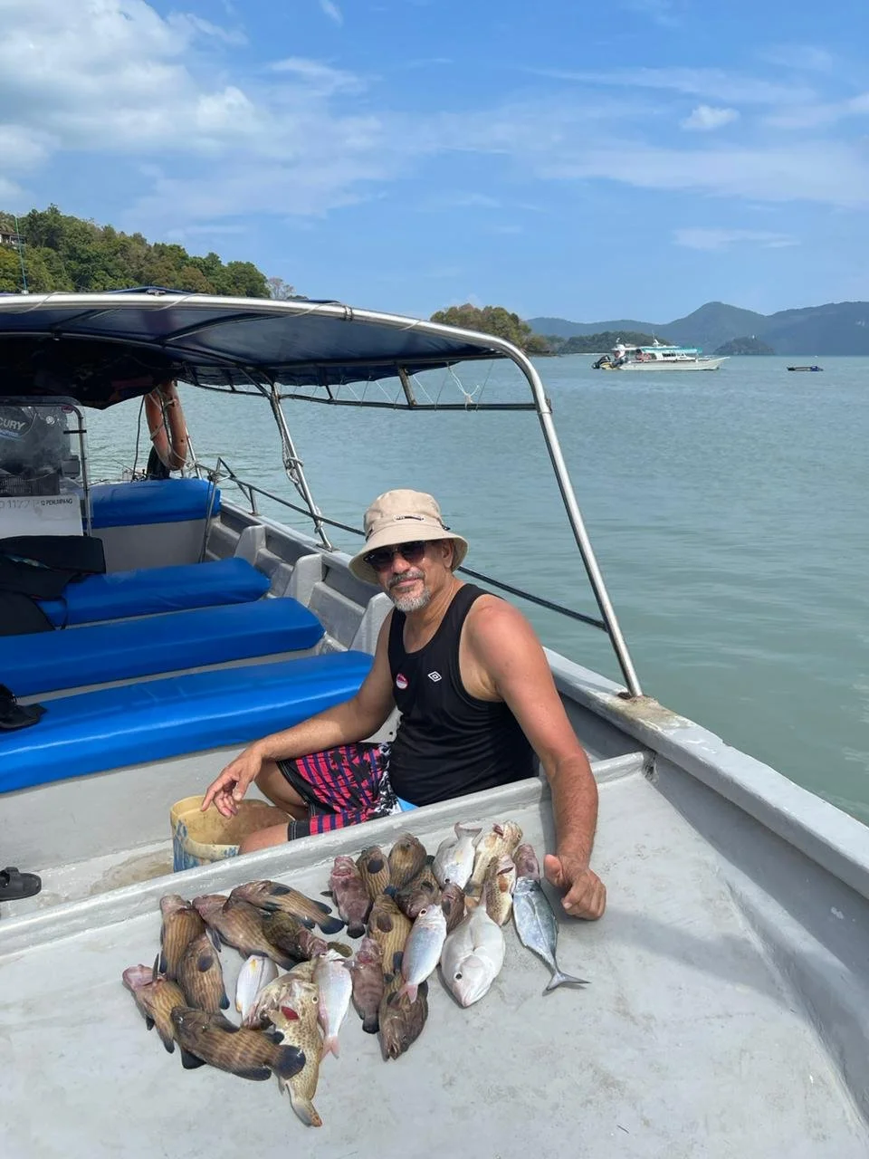 Man sitting on a boat with a display of caught fish, wearing sunglasses, a hat, a black tank top, and patterned shorts, with a shoreline, water, and boats in the background.