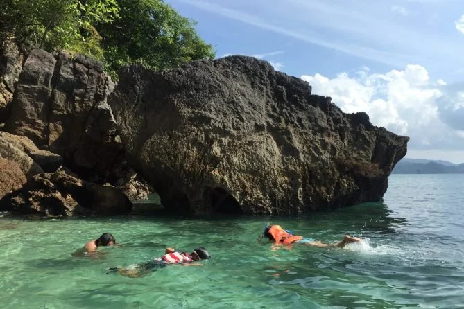 People swimming near large rocks and a shoreline with trees under a partly cloudy sky.