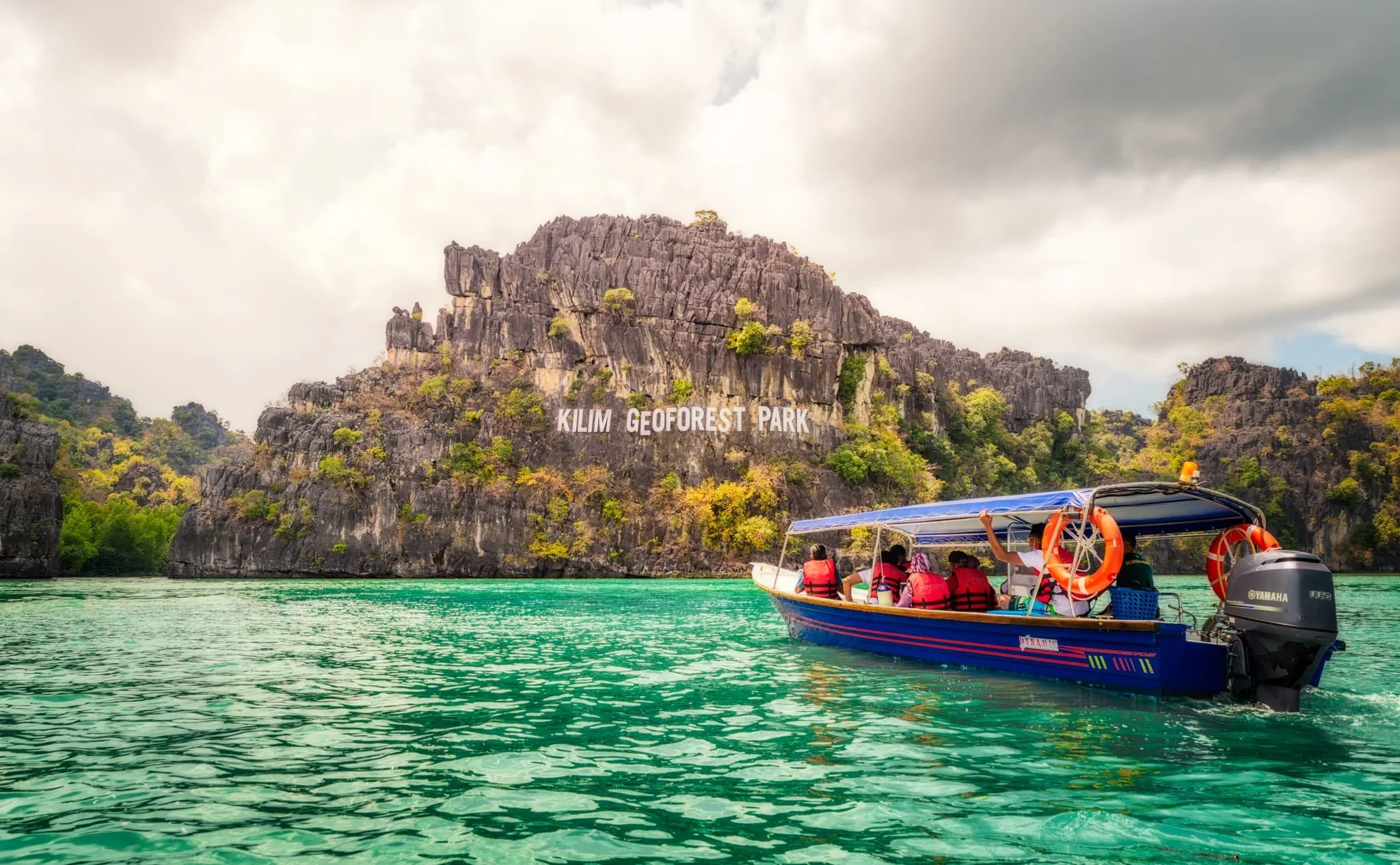 A boat with passengers wearing life jackets on a body of water near a rocky island with a sign that reads 'Kilim Geoforest Park' in Malaysia.