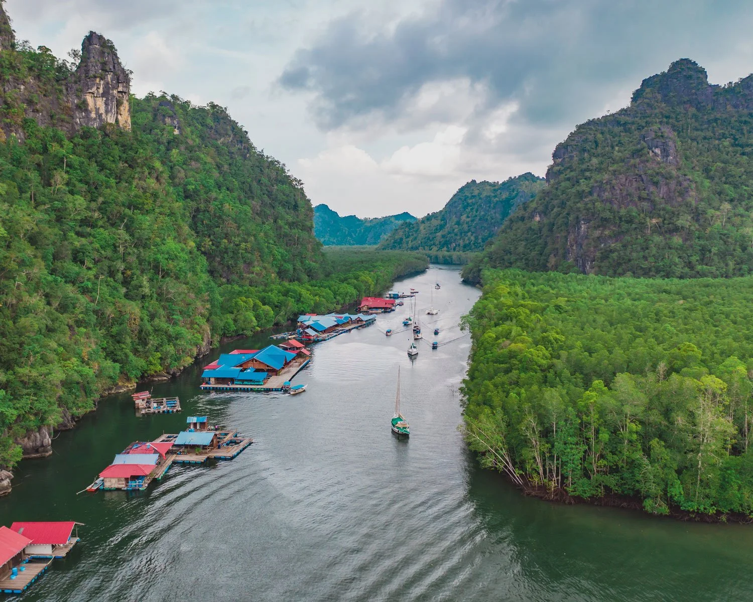 A river through a green valley with mountains on both sides, some boats on the water, and structures with blue and red roofs along the riverbank.