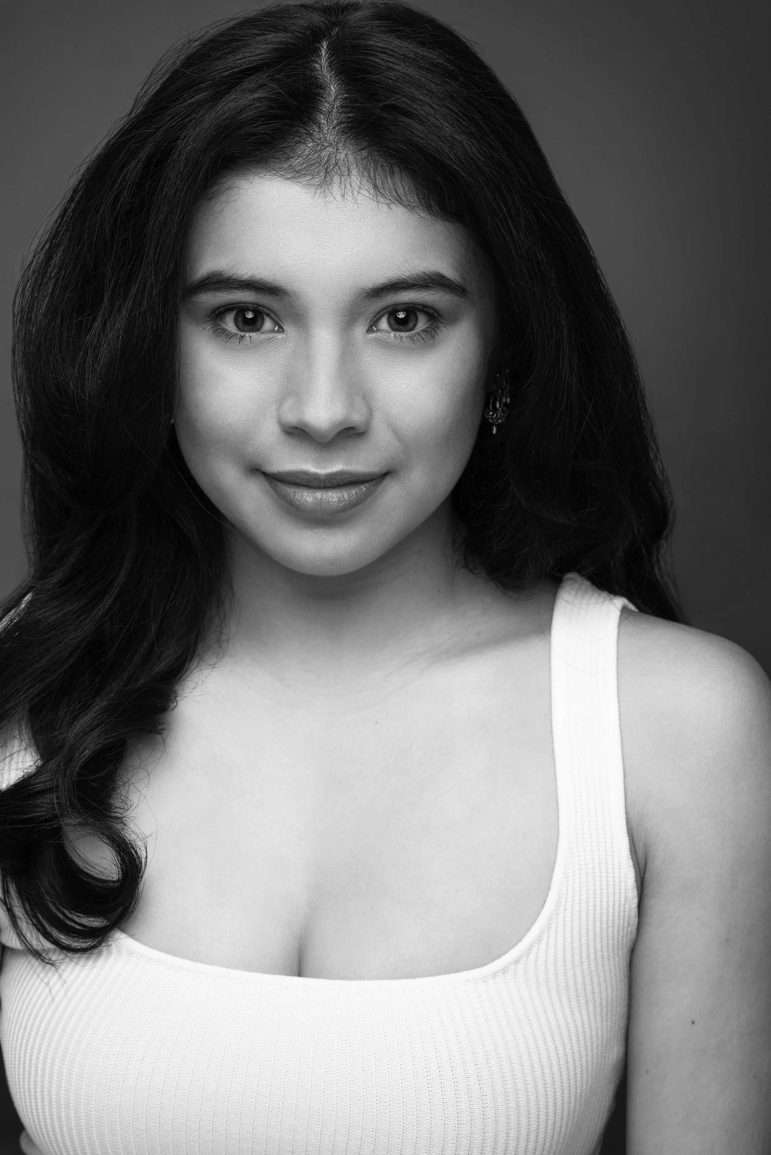 Black and white portrait of a young woman with long dark hair, smiling softly, wearing a sleeveless top, earrings, and looking directly at the camera.