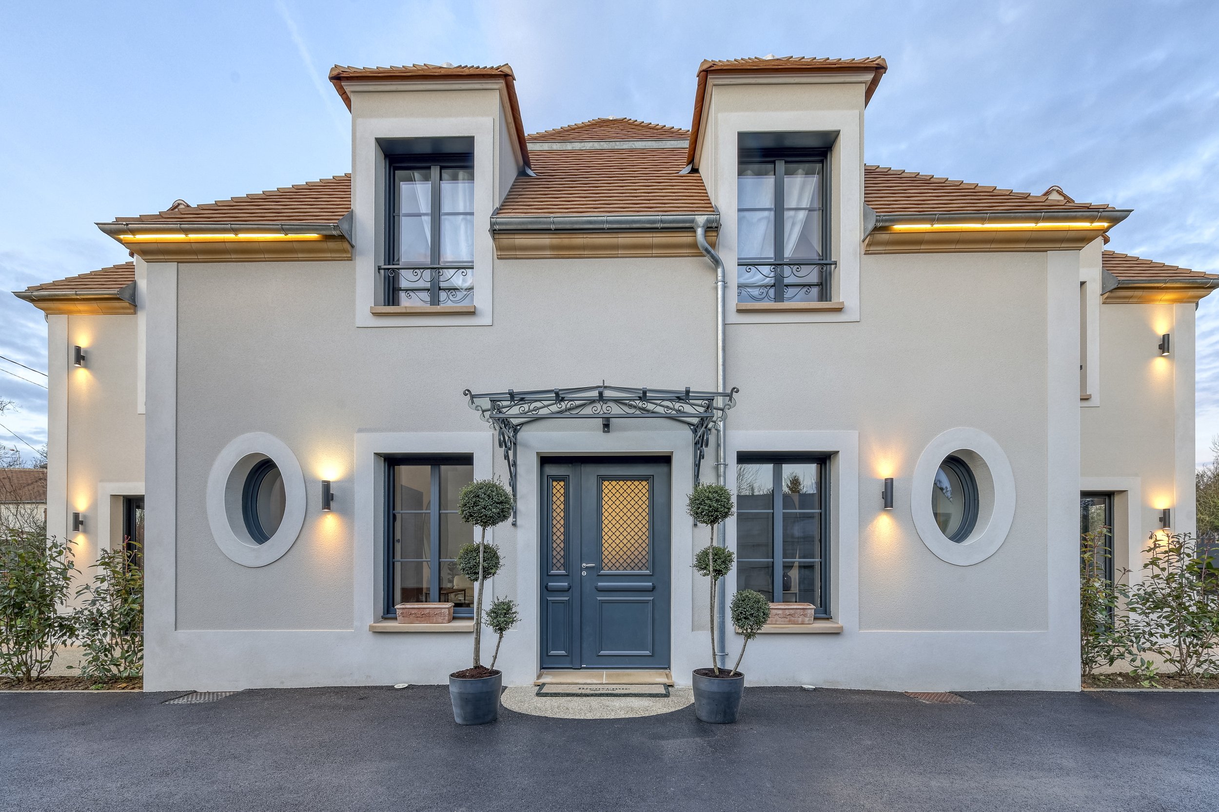 Maison moderne à deux étages avec façade blanche, fenêtres rondes et rectangulaires, porte bleue centrale, jardins avec plantes taillées en forme de boule, éclairage extérieur.