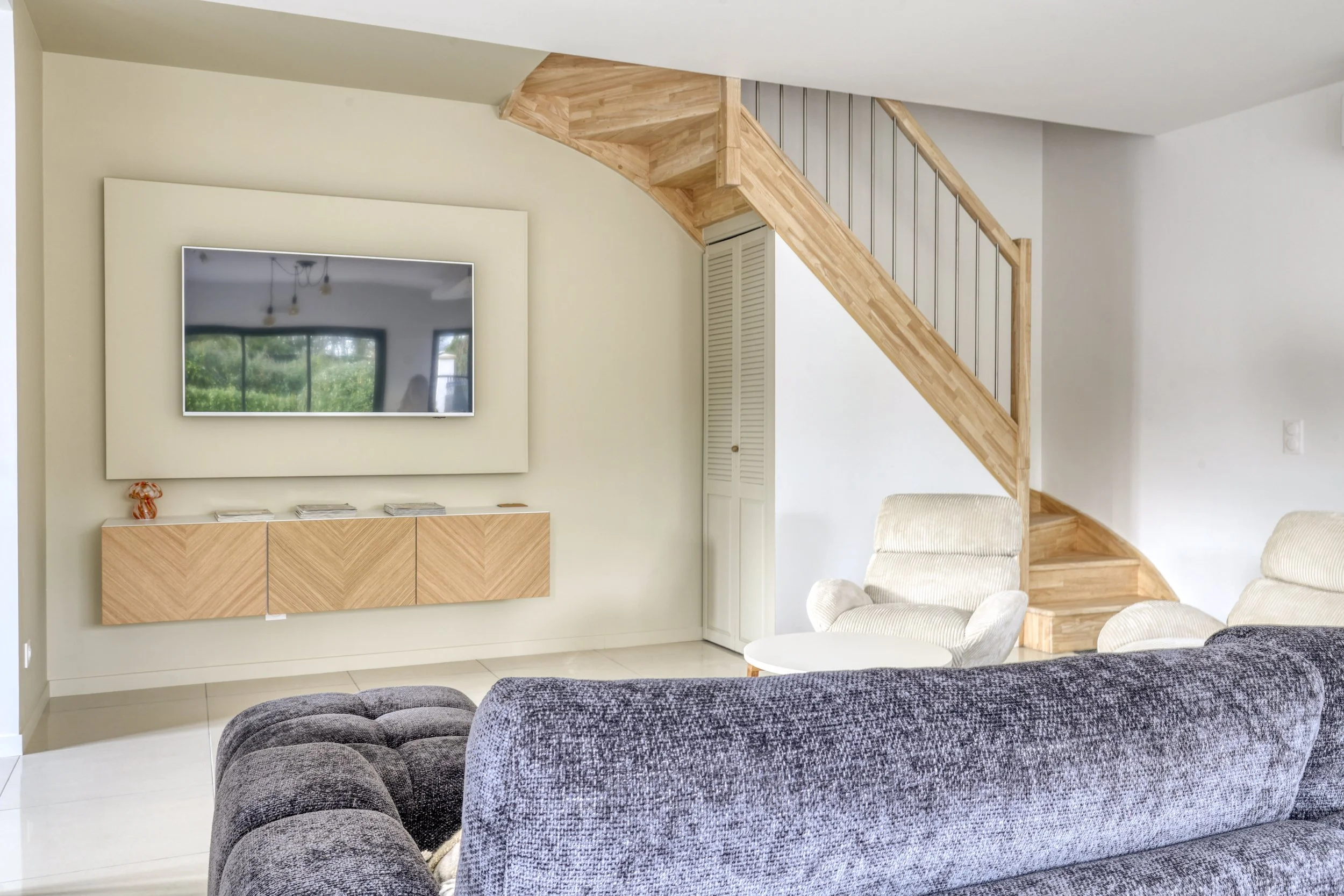 Salon moderne avec canapé gris, fauteuils beige, télévision à l wall, escalier en bois et rangement en bois