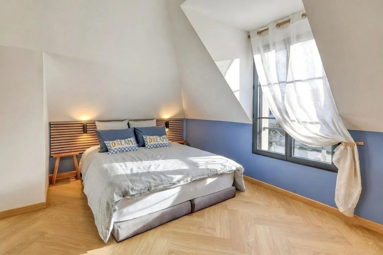 Chambre à coucher lumineuse avec un lit double, draps blancs, et des coussins bleus. La chambre a un mur bleu et une grande fenêtre avec des rideaux blancs, laissant entrer la lumière naturelle.