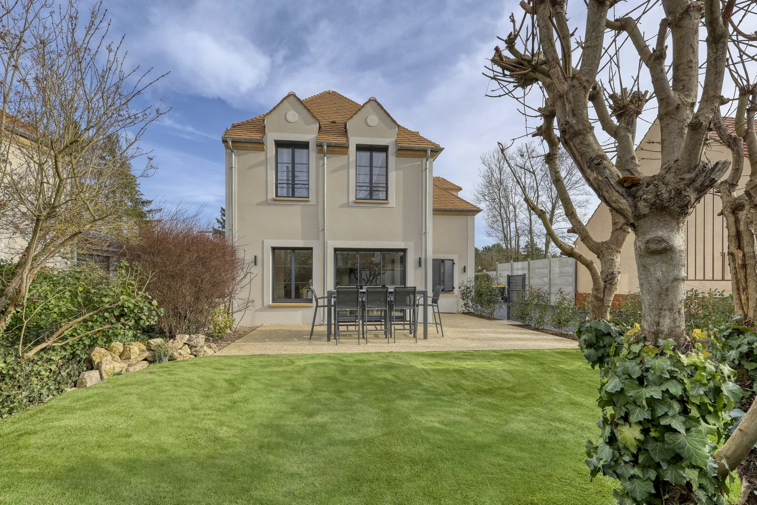 Jardin de maison avec pelouse, arbres et terrasse avec table et chaises, maison à deux étages avec fenêtres