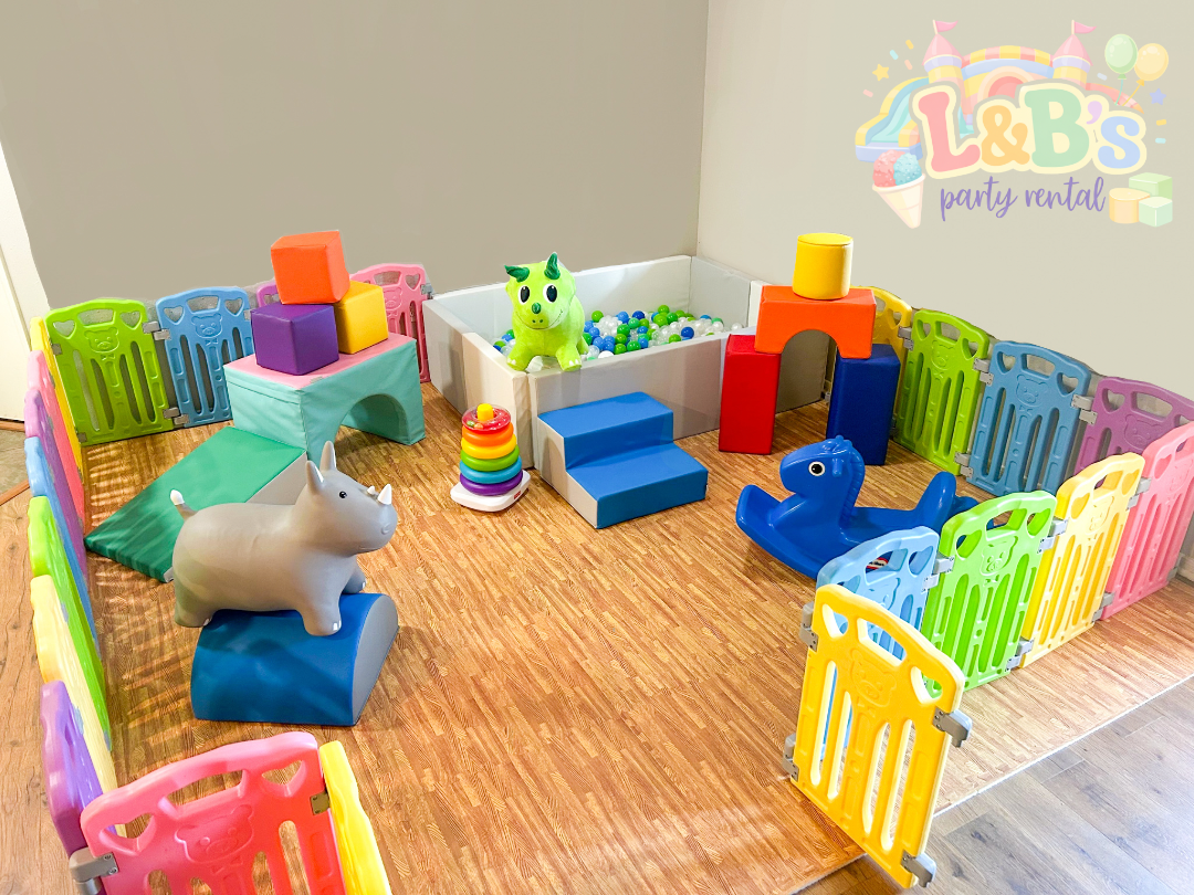 Indoor children's play area with colorful plastic fences, animal-shaped soft seating, stacking rings, foam mats, and a ball pit with a unicorn plush toy inside.