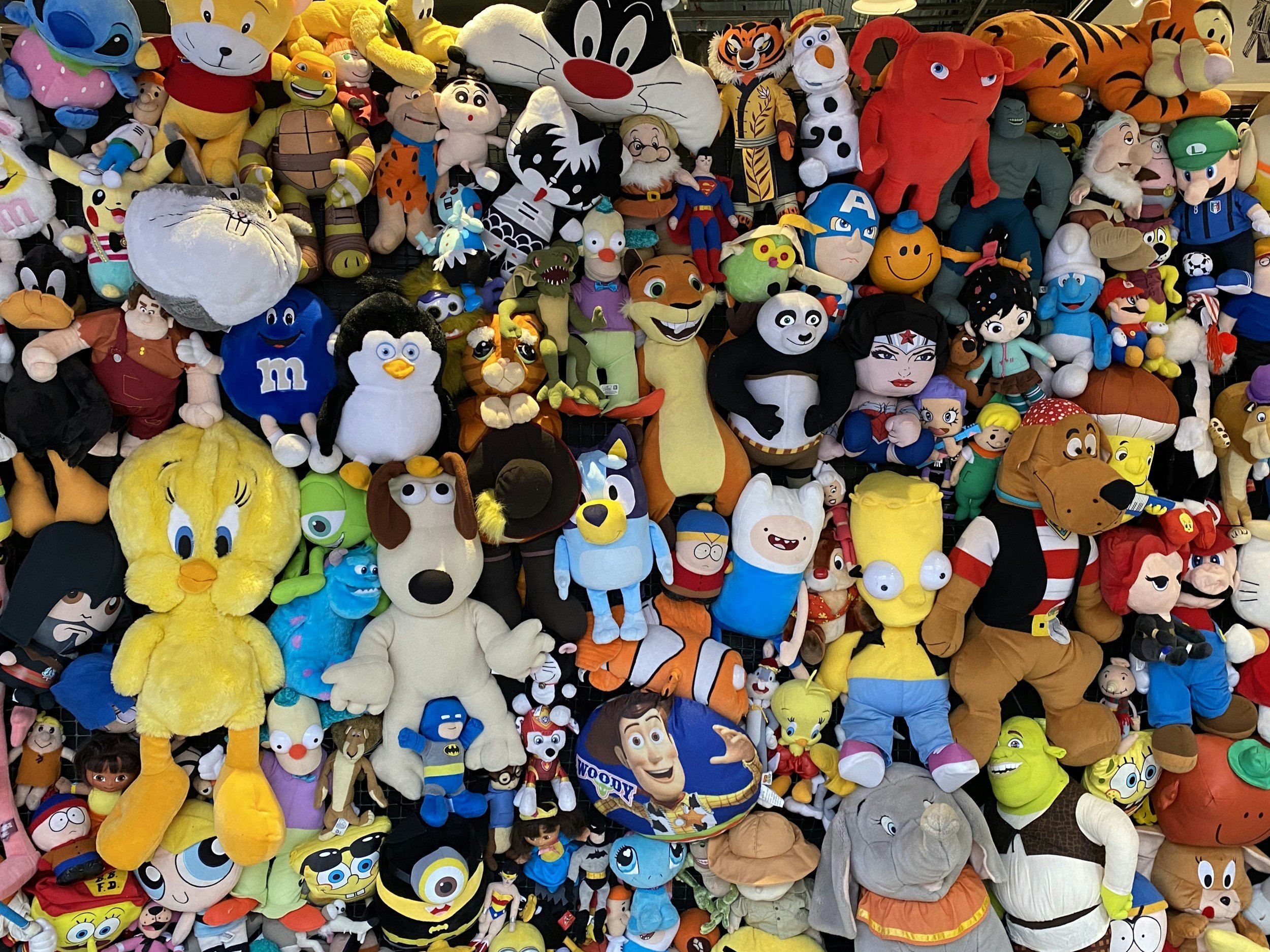 A collection of colorful plush toys featuring various characters from animated movies, TV shows, and comic books, including Scooby-Doo, Wonder Woman, Shrek, Minions, and other popular animated characters.