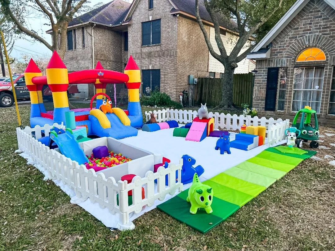 An outdoor children's play area with a white picket fence, inflatable bounce house, climbing toys, a ball pit, and ride-on toys set up on the grass outside houses.