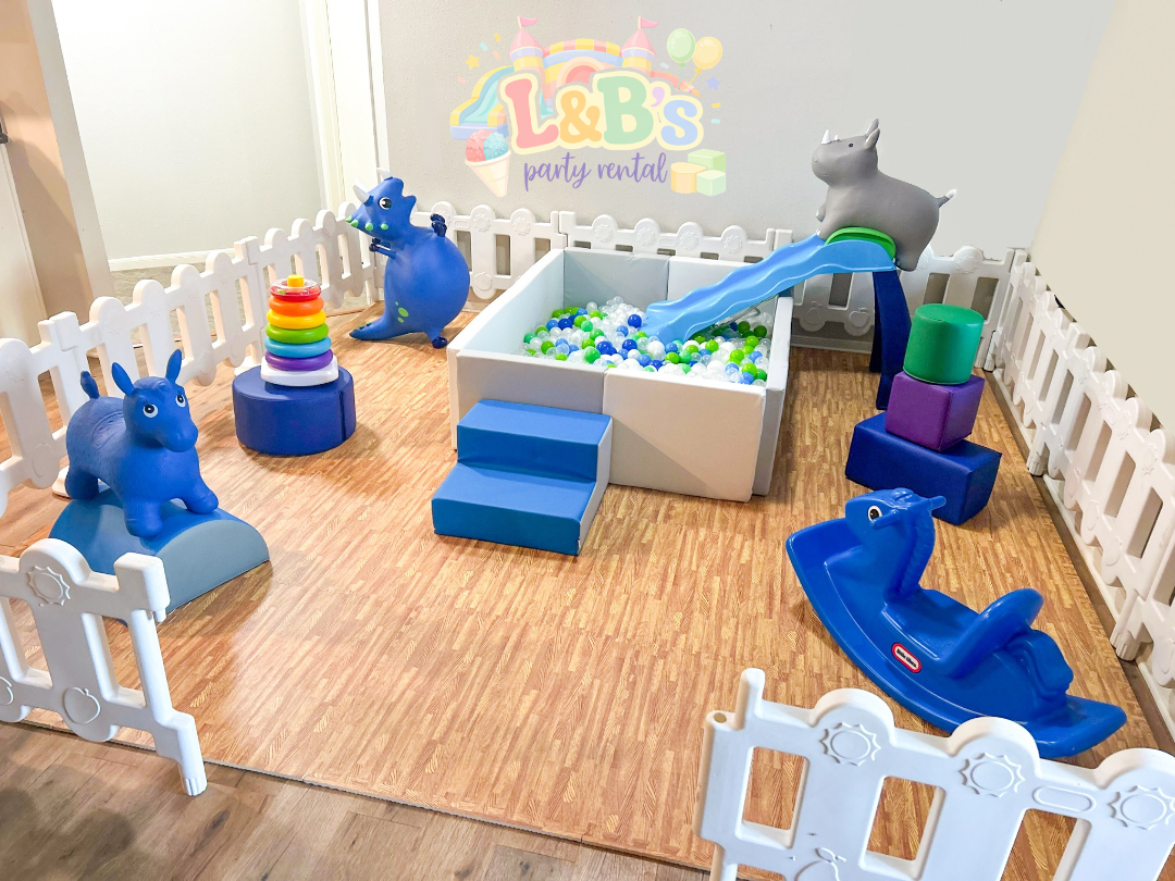 Indoor children's play area with a ball pit, plastic animals, a slide, and soft blocks, enclosed by white plastic fence panels, with a sign reading "L&B's party rental" on the wall.