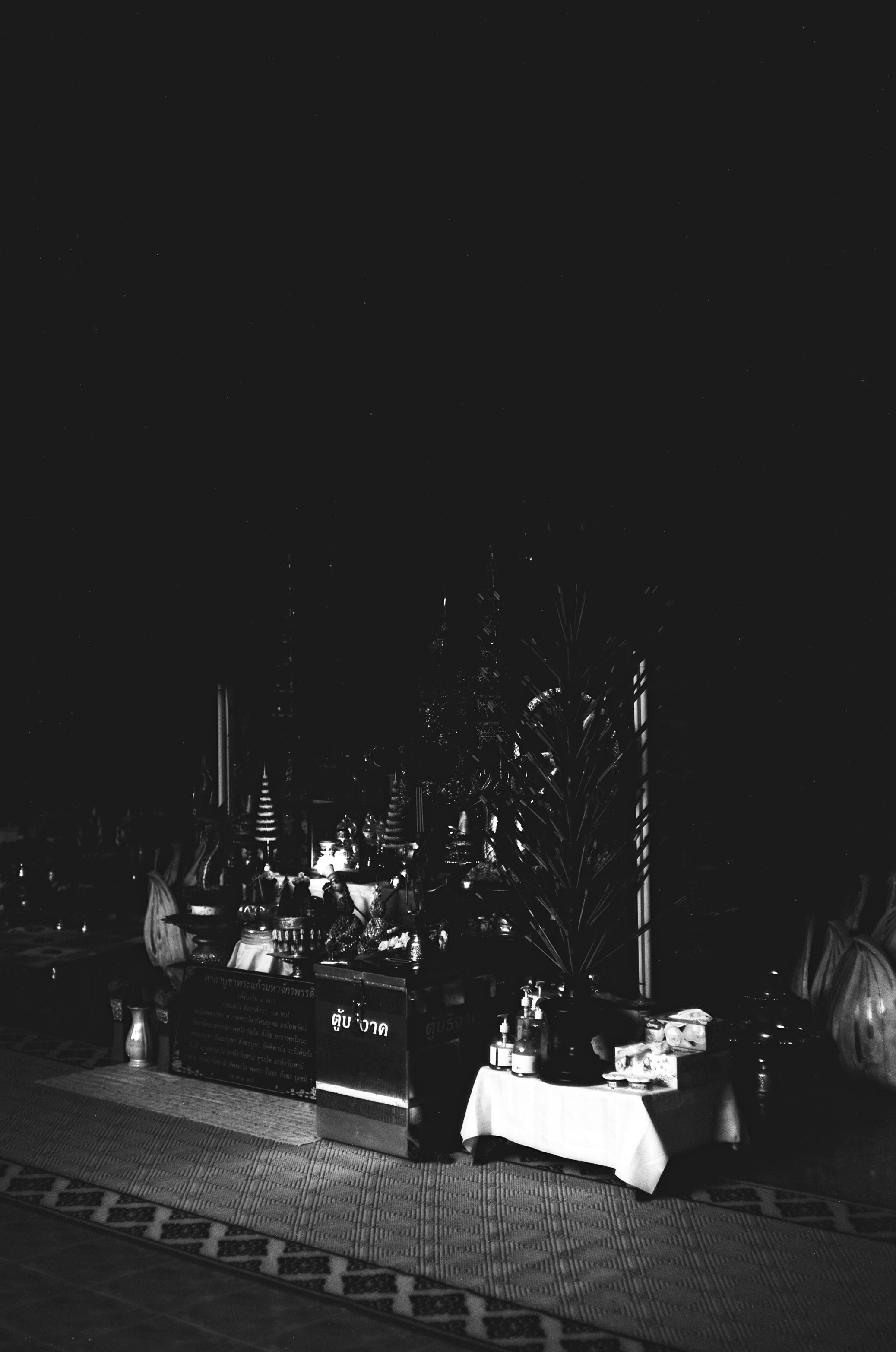 A black and white photo of a night scene with attach tables and objects, including bottles and containers, against a backdrop of traditional Thai decorations and lanterns.