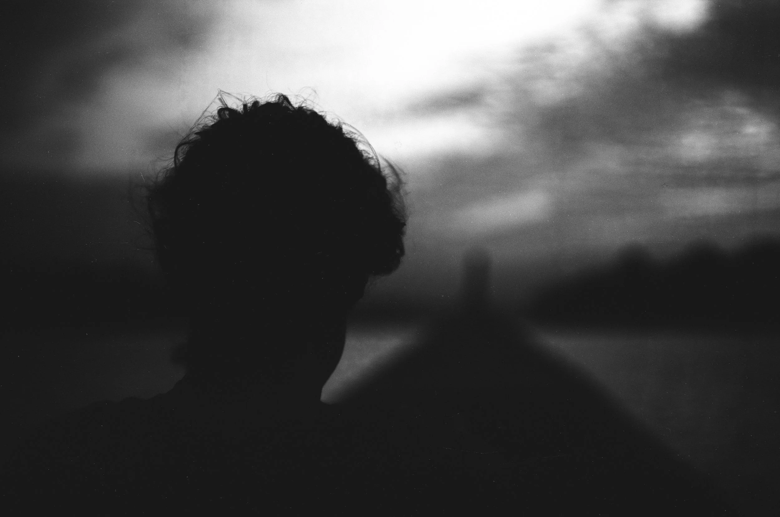 Silhouette of a person with curly hair standing outdoors during dusk or dawn, with a cloudy sky and a blurry background.