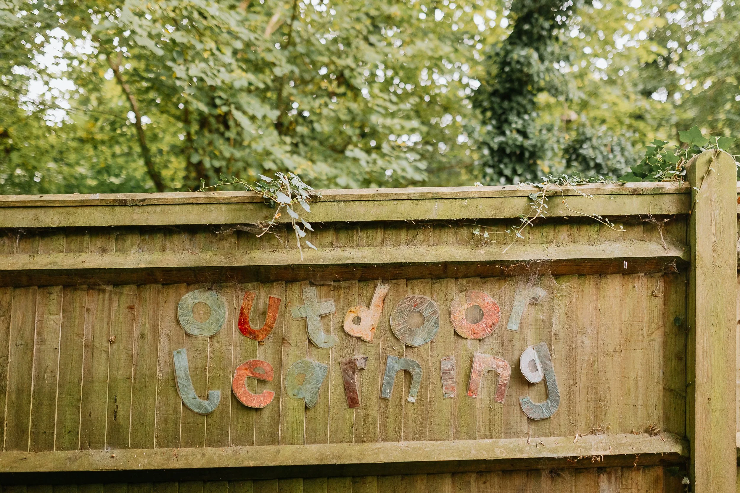 A wooden fence with colorful metal letters spelling 'outdoor' hanging on it, with overgrown green foliage and trees in the background.