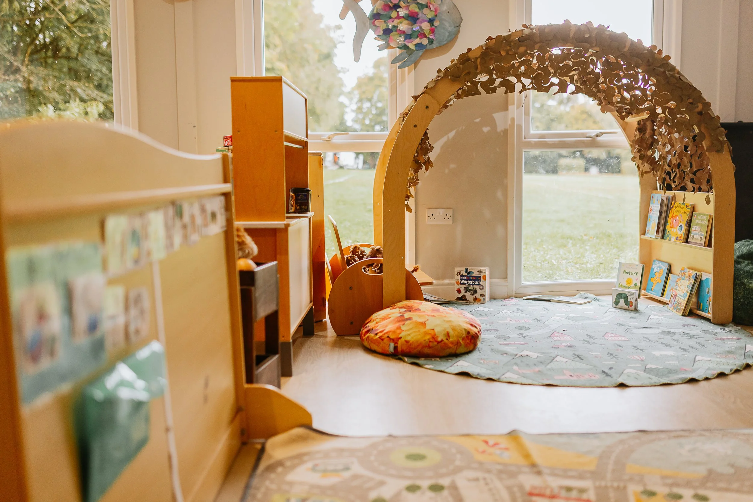 A cozy children's reading nook with a large window and natural light. There are children's books displayed on wooden shelves, a colorful round floor cushion, and a decorative arc with leaf patterns. Outside, trees and a field are visible through the window.