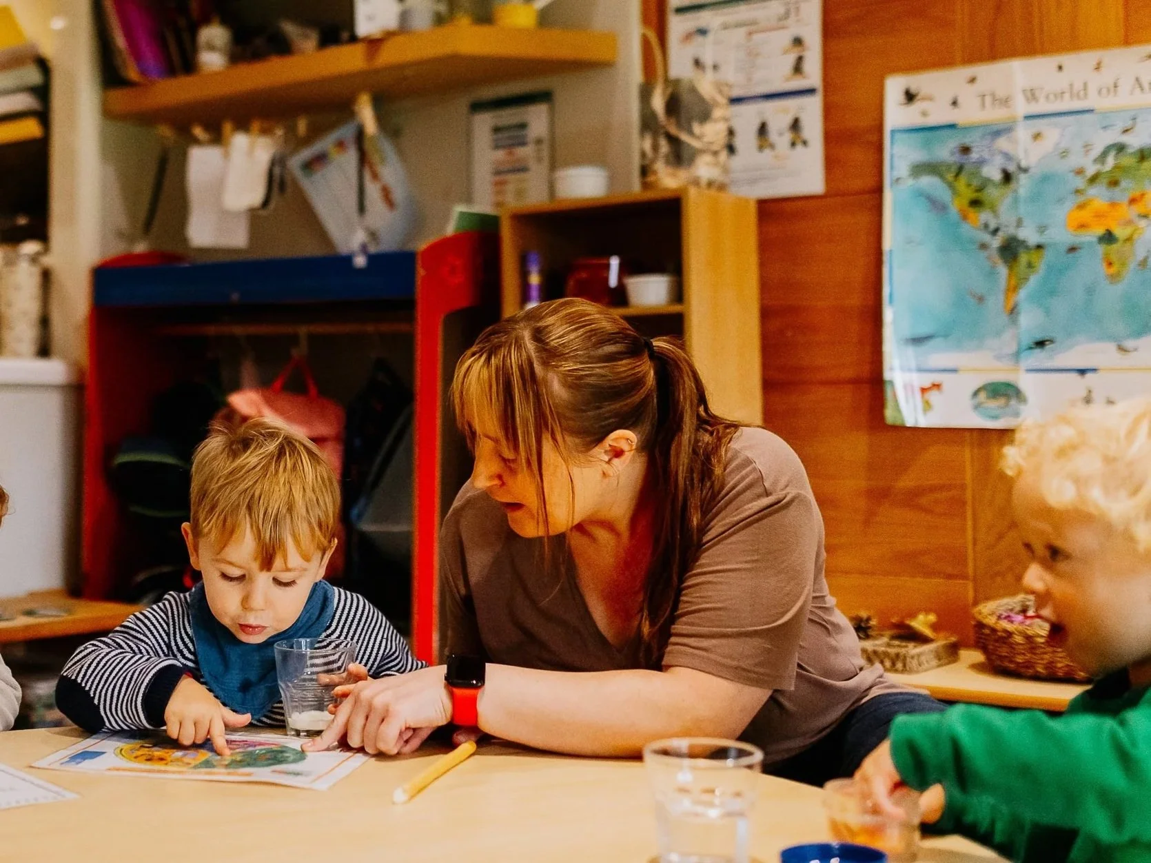A woman sitting at a table with two young children, one boy and one girl, in a classroom setting. The woman is leaning towards the children, who are engaged in an activity with papers and a glass of water. The classroom has educational posters on the wall and shelves with supplies.